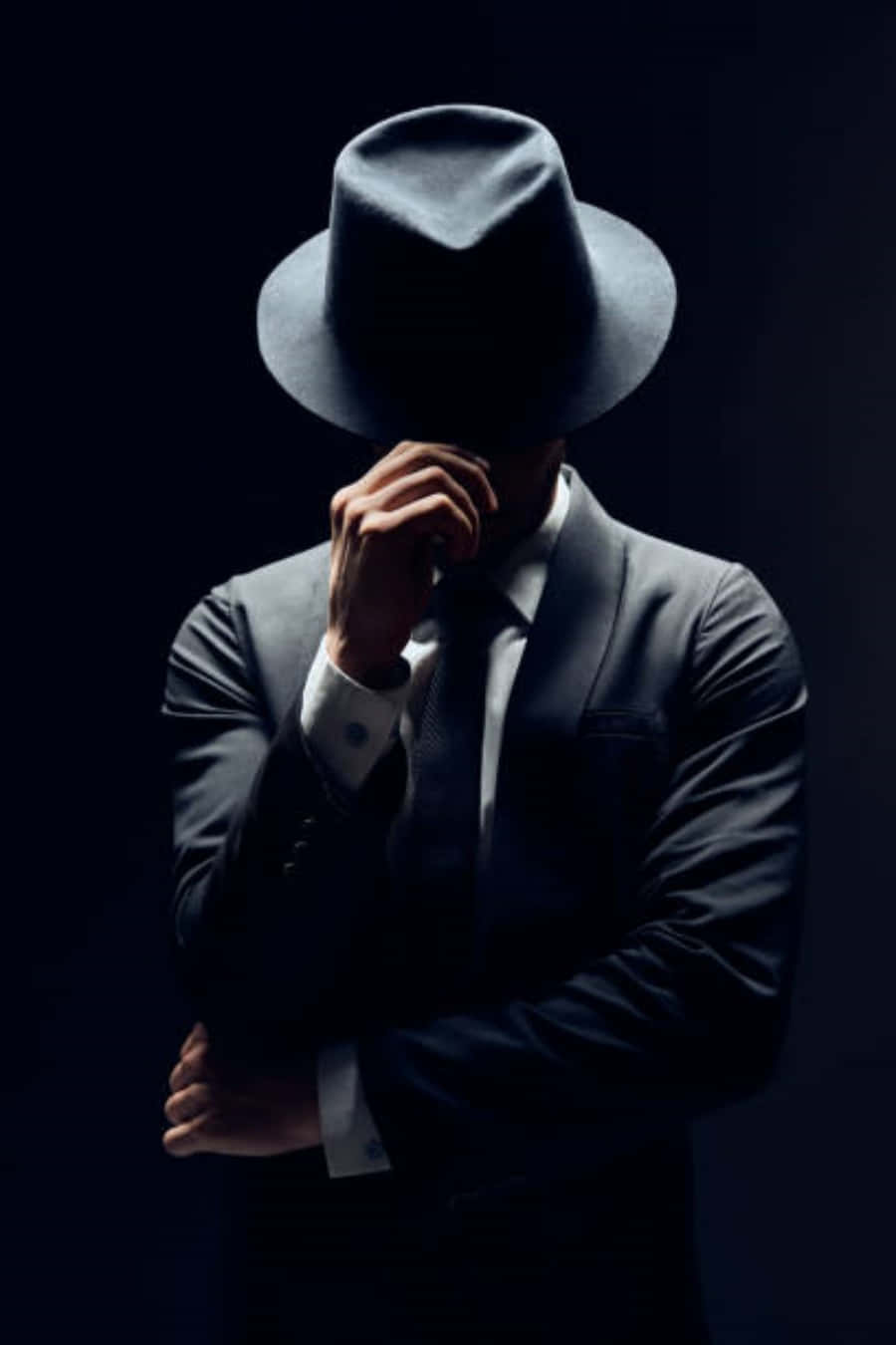 Mafia 桌布 壁紙 — 一個神秘的男人穿著深色西裝和軟呢帽，在黑色背景前用手遮住臉。