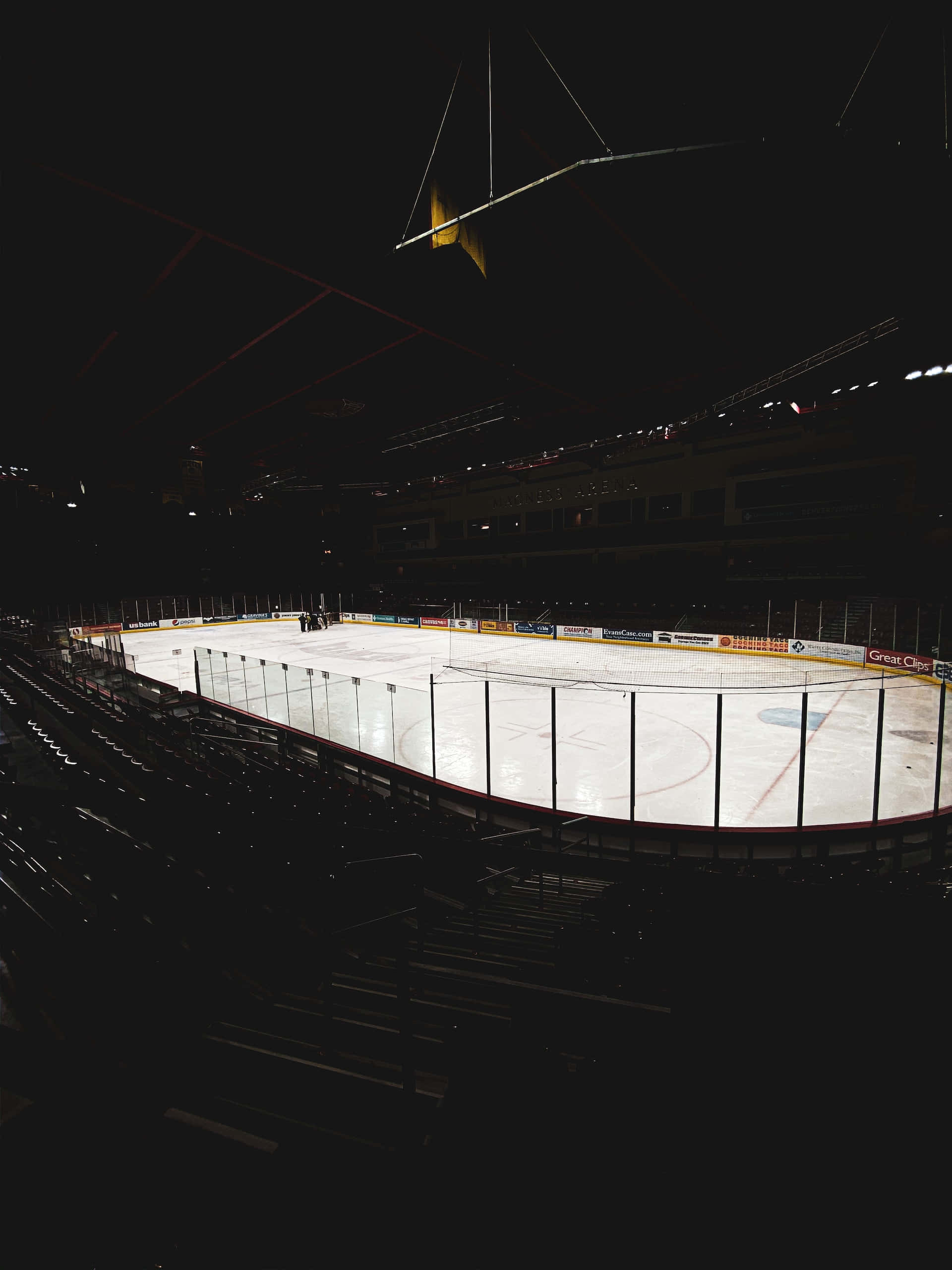 Ice Rink 桌布 壁紙 — 一個空置的冰球場，由頭頂燈光照明，位於黑暗的場館內，周圍是一排排空座位。