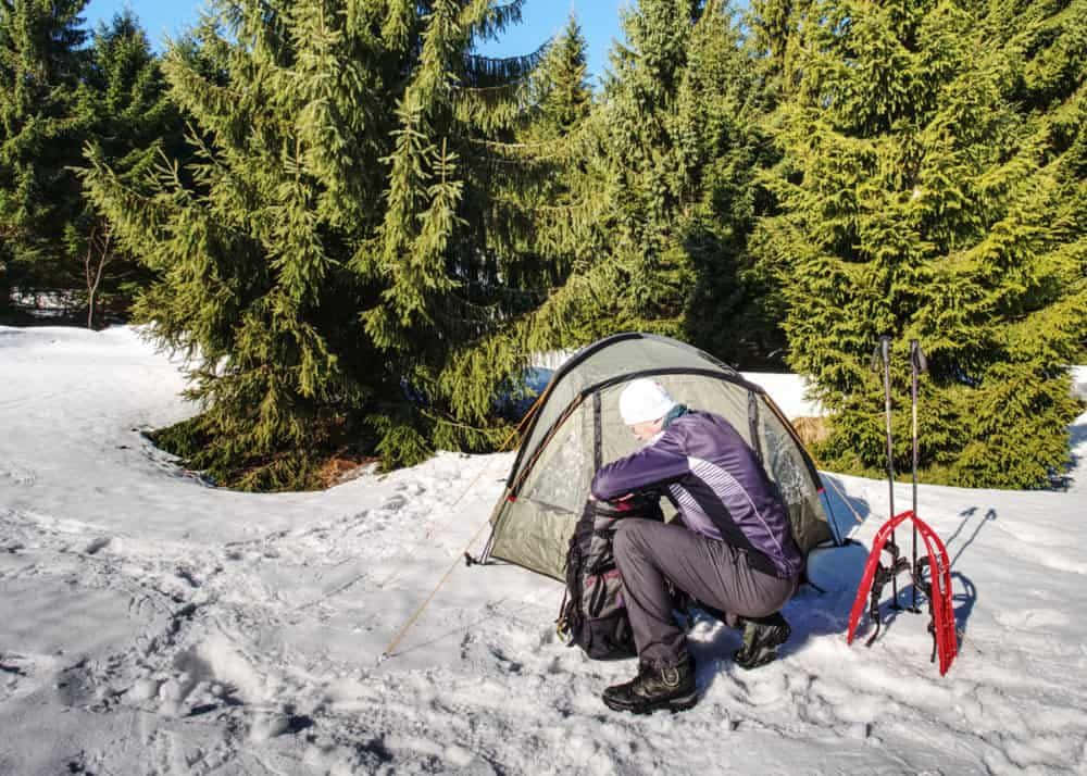 Winter Camping 桌布 壁紙 — 一個穿著冬季裝備的人正在陽光明媚的雪地森林中搭建帳篷，周圍是常青樹。