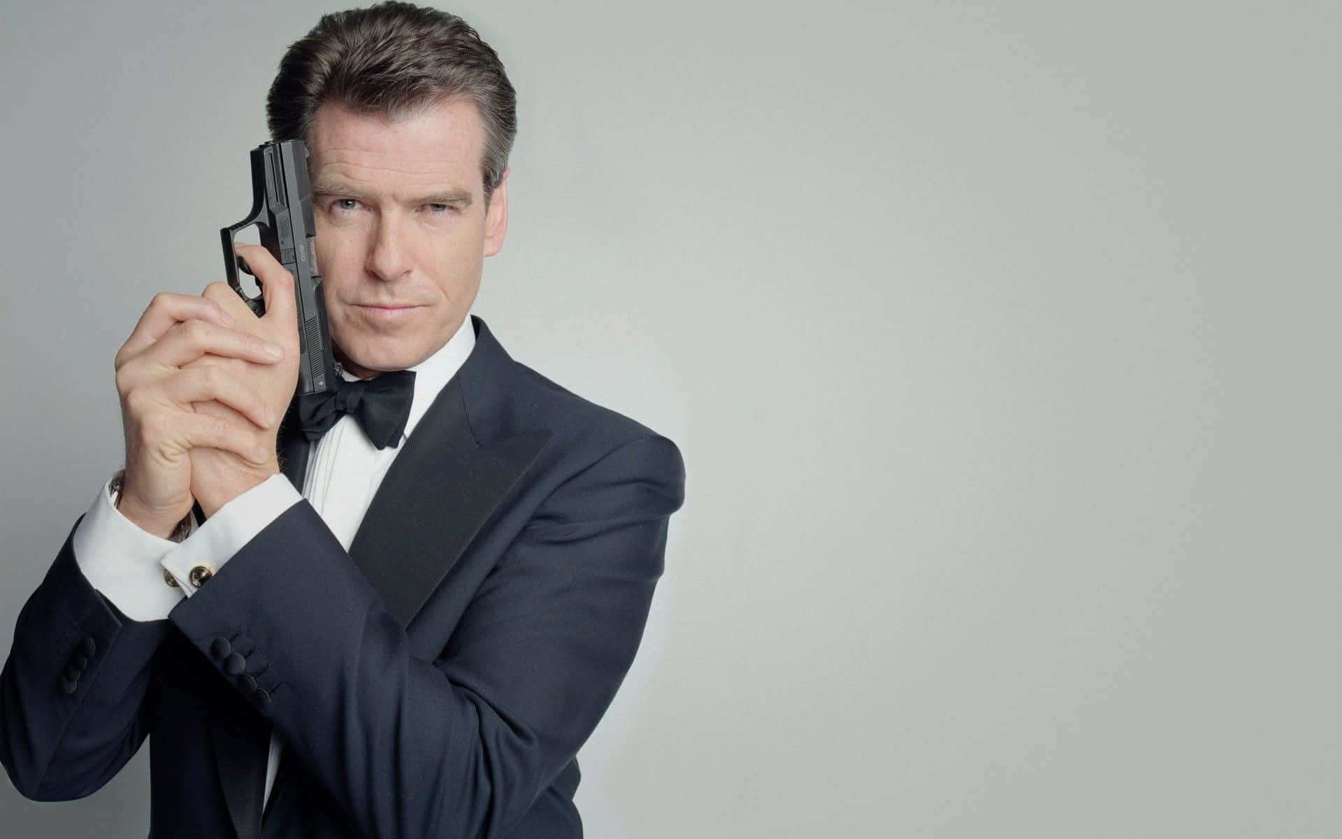 pierce brosnan 桌布 壁紙 — 一個穿著利落黑色燕尾服的男人，在純灰色背景下，手持手槍，神情嚴肅。
