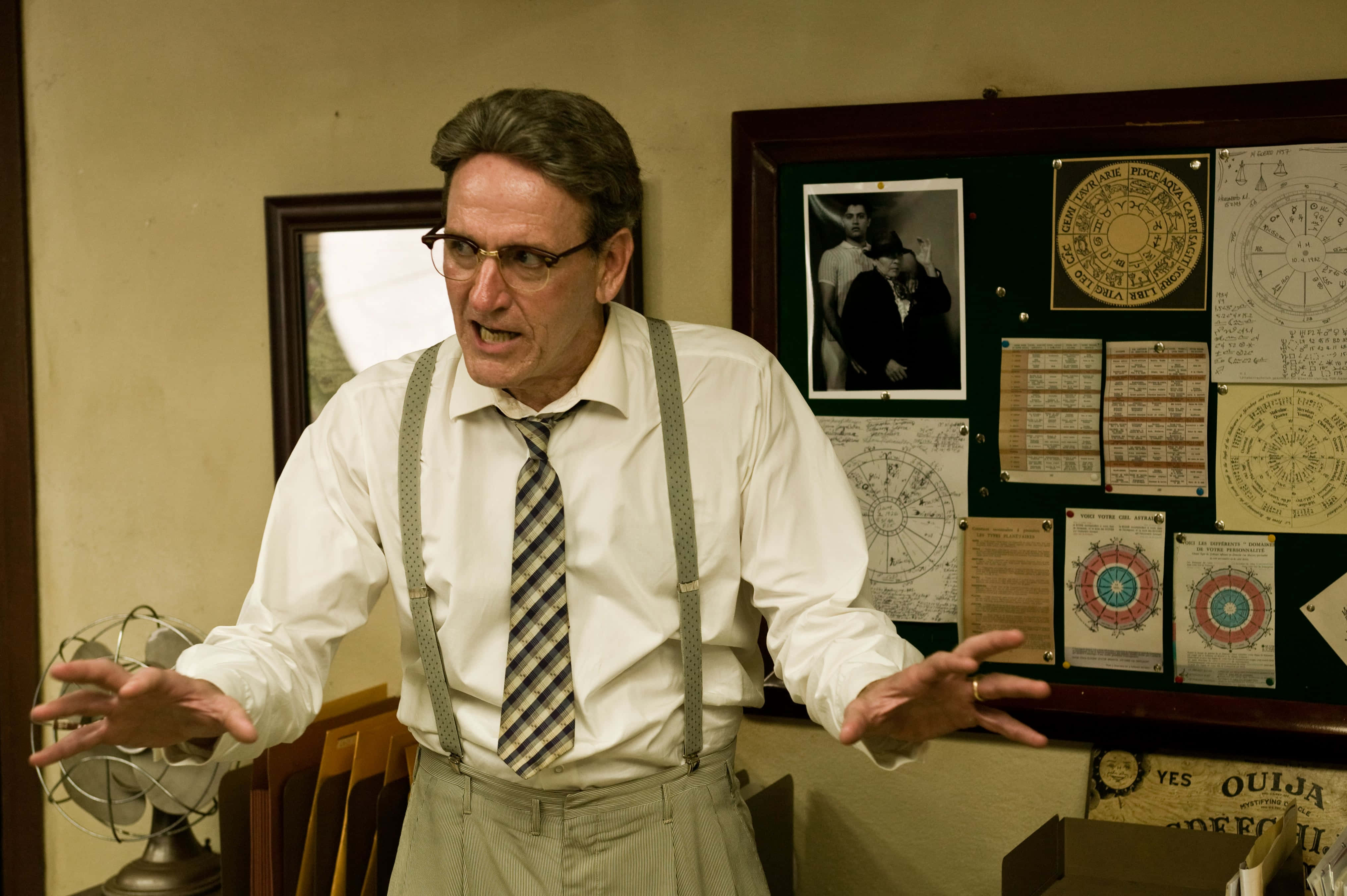 Richard Jenkins 桌布 壁紙 — 一個穿著吊帶褲和眼鏡的男人在一個雜亂的辦公室裡誇張地做手勢，辦公室裡充滿了神秘海報和一個通靈板。