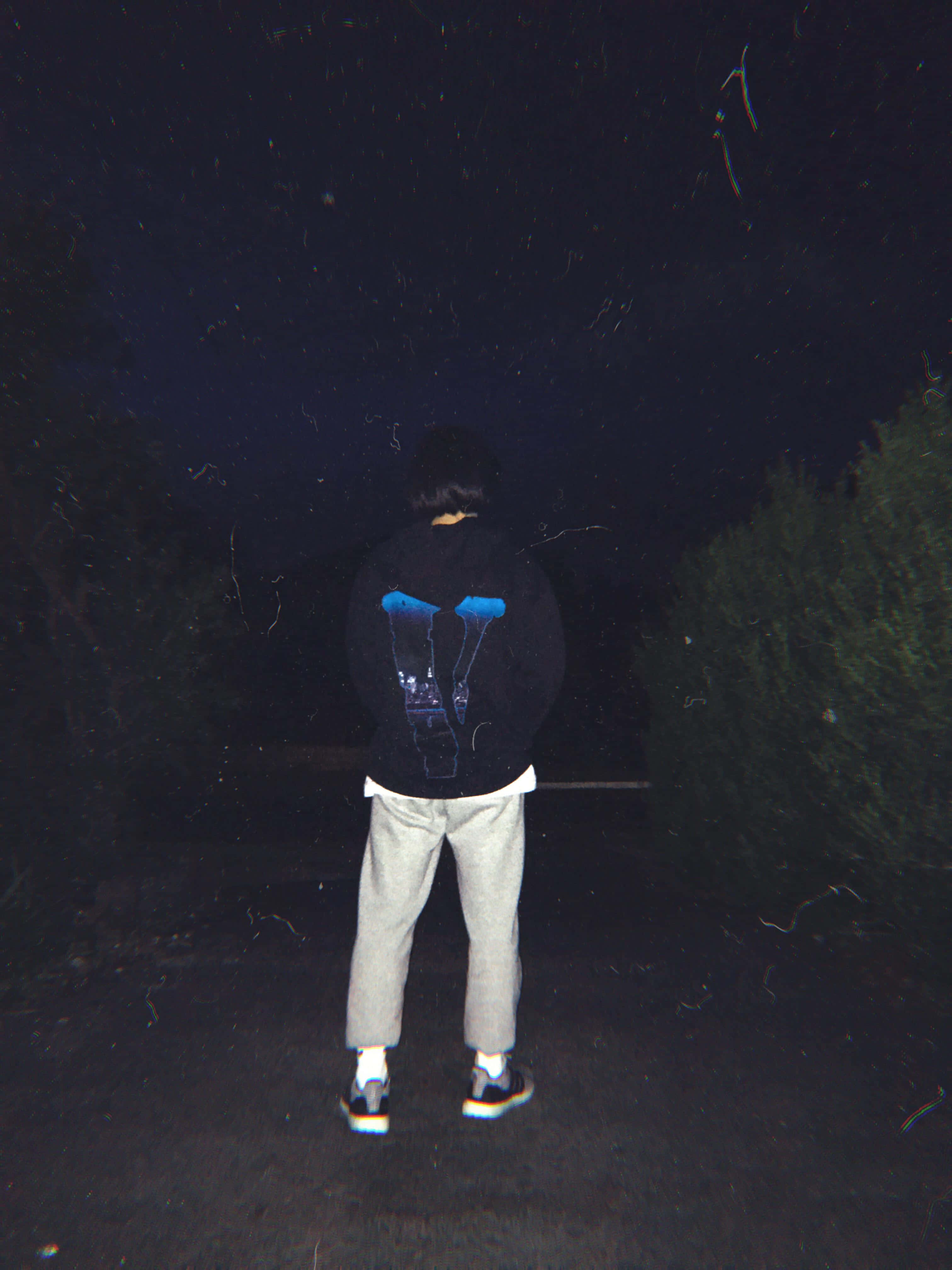Vlone 桌布 壁紙 — 一個穿著帶有藍色圖案的黑色連帽衫的人，在夜晚獨自站在星空下，周圍是黑暗的樹木。