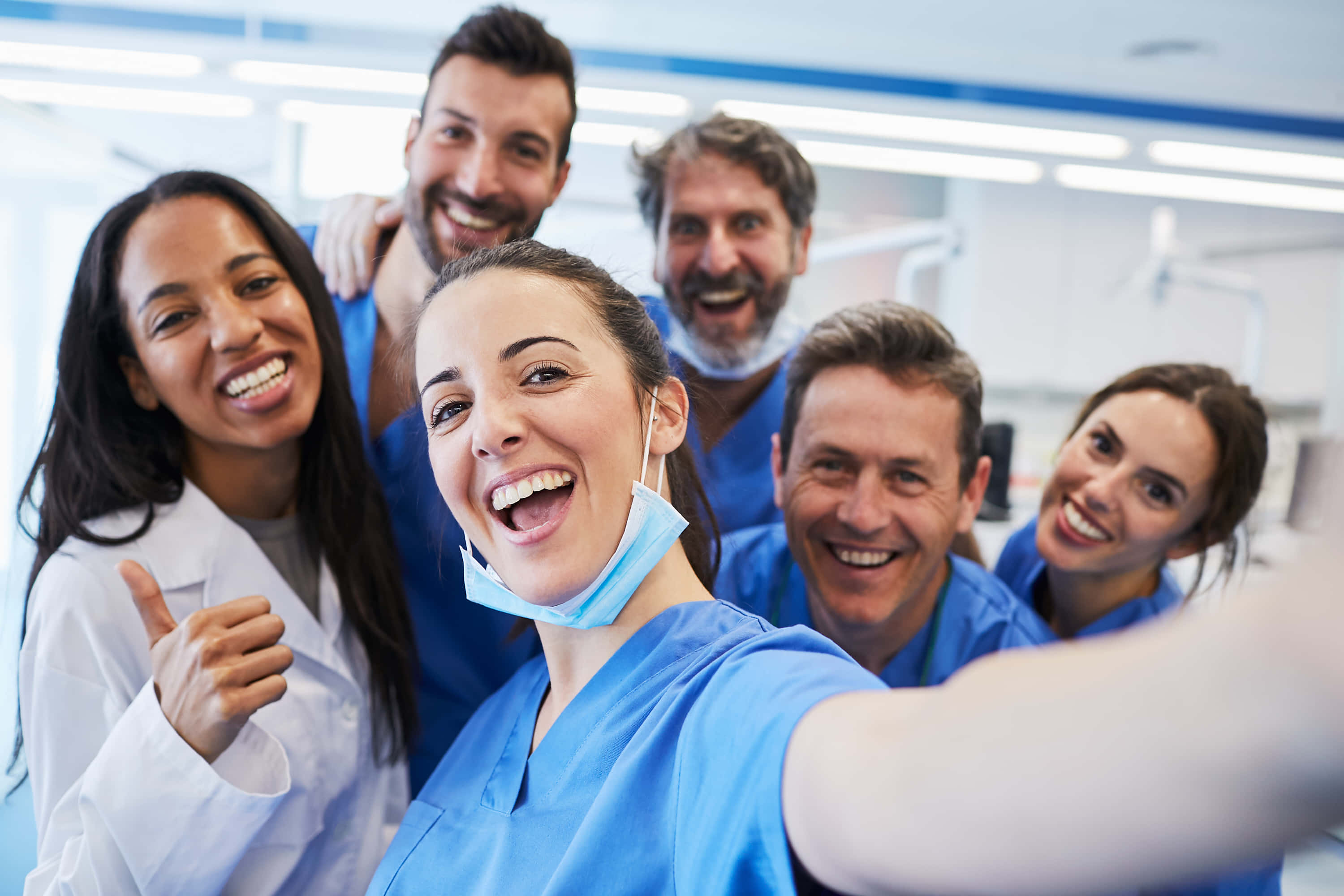 Nursing 桌布 壁紙 — 一個穿著手術服和實驗室外套的歡樂醫療團隊在明亮的臨床環境中微笑自拍。