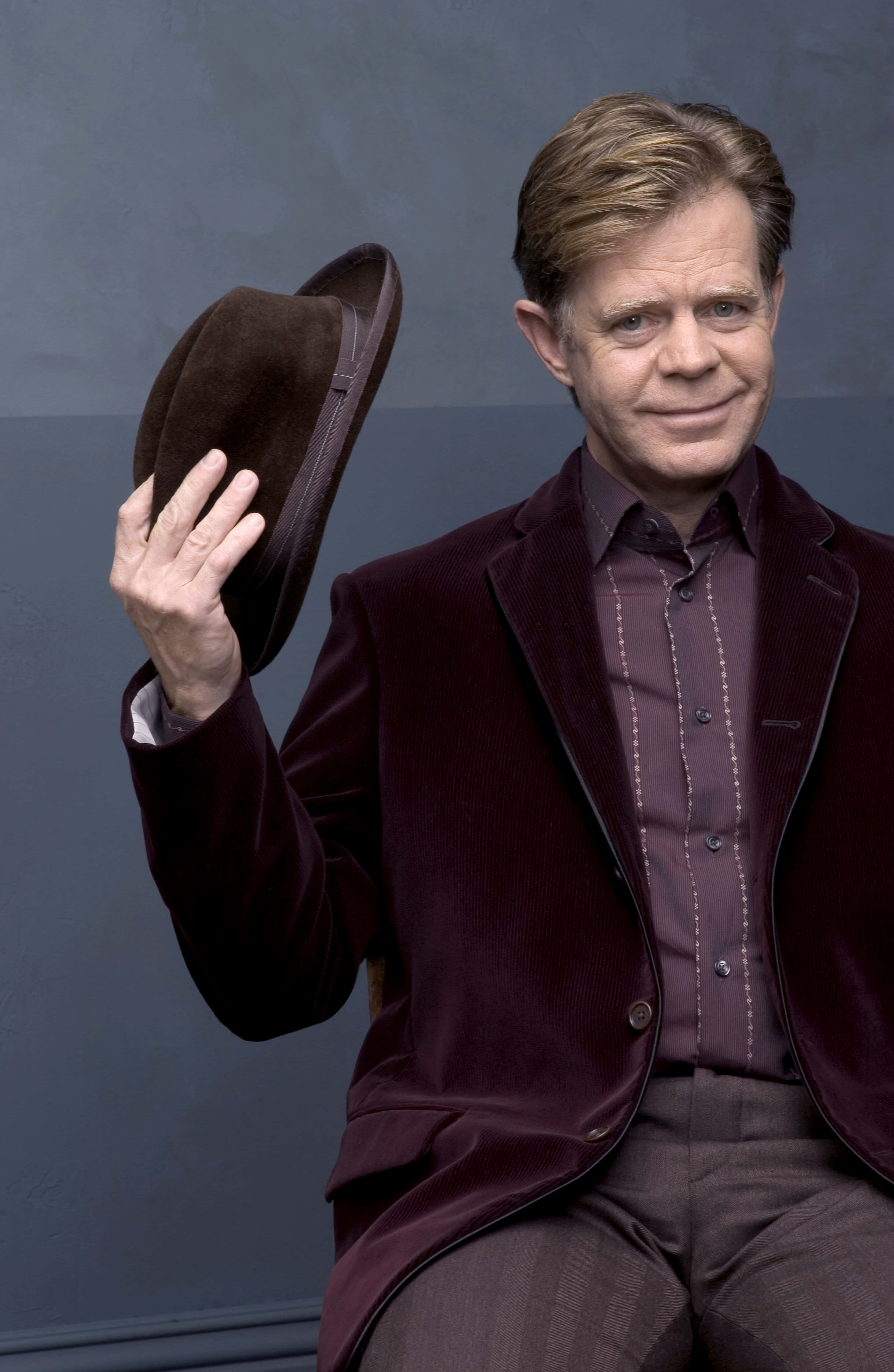 william h. macy 桌布 壁紙 — 一個穿著栗色天鵝絨西裝的男人，坐在柔和的灰色背景前，手持一頂棕色帽子。