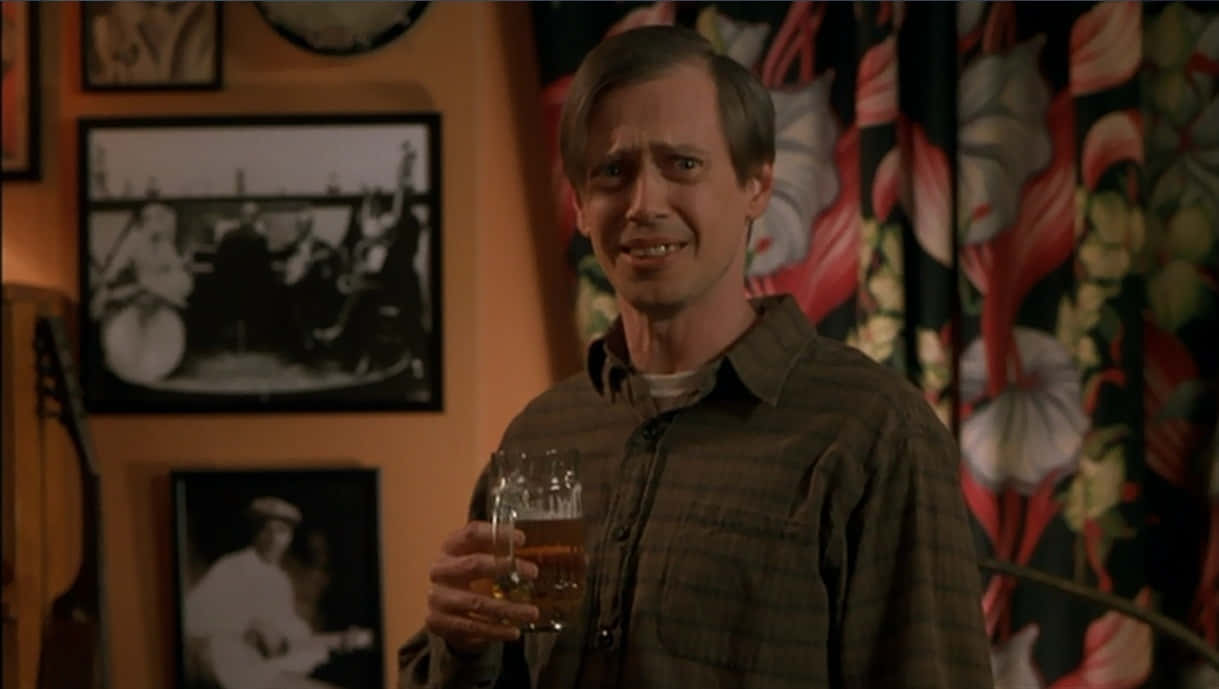 steve buscemi 桌布 壁紙 — 一個穿著格仔恤衫嘅男人喺燈光溫暖嘅房間入面，手拎住啤酒杯，房間有花卉圖案窗簾同埋牆上嘅相框。