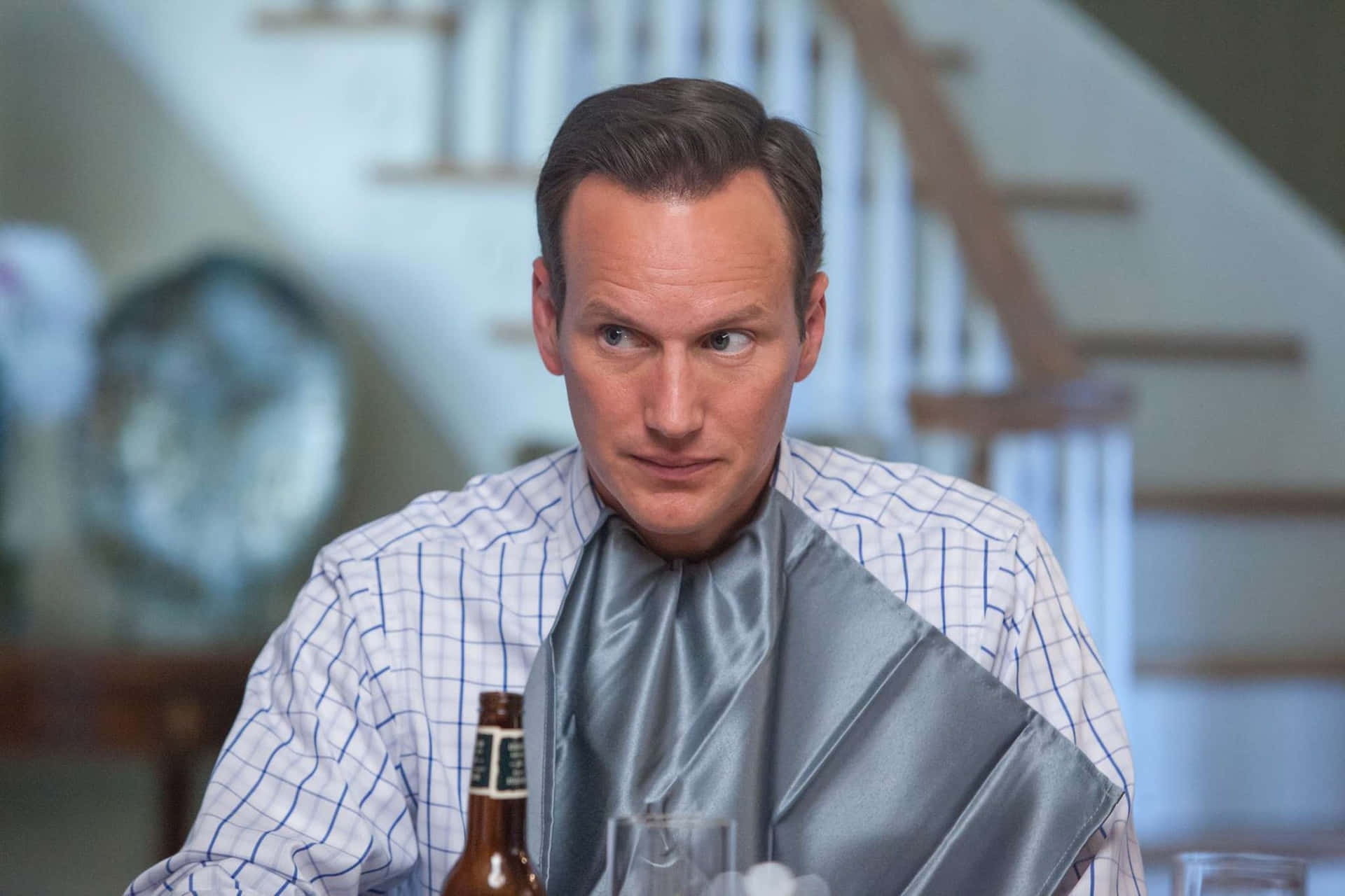 Patrick Wilson 桌布 壁紙 — 一個穿著格仔恤衫的男人坐在桌旁，肩上搭著一條銀色餐巾，旁邊放著一個啤酒瓶，表情嚴肅地望向鏡頭外。