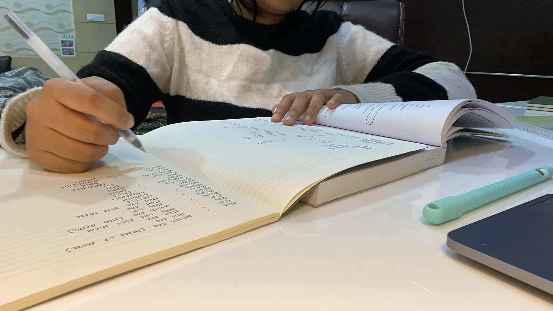 溫習 桌布 壁紙 — 一個穿著條紋毛衣的人正在白色桌子上用筆記本電腦和綠色筆在筆記本上寫字。