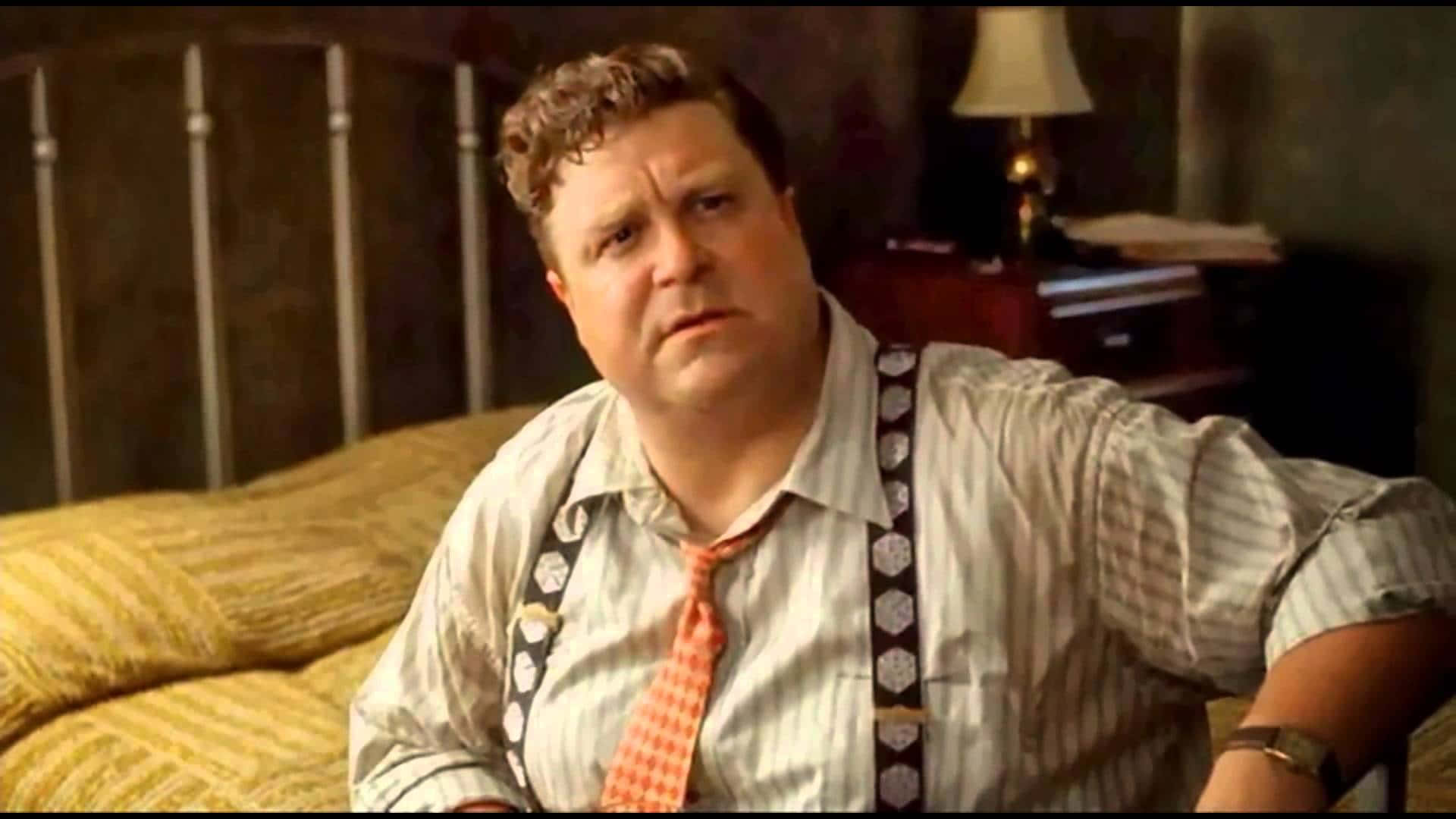 John Goodman 桌布 壁紙 — 一個穿著條紋襯衫和吊帶的男人坐在黃色床上，表情困惑，房間光線昏暗。