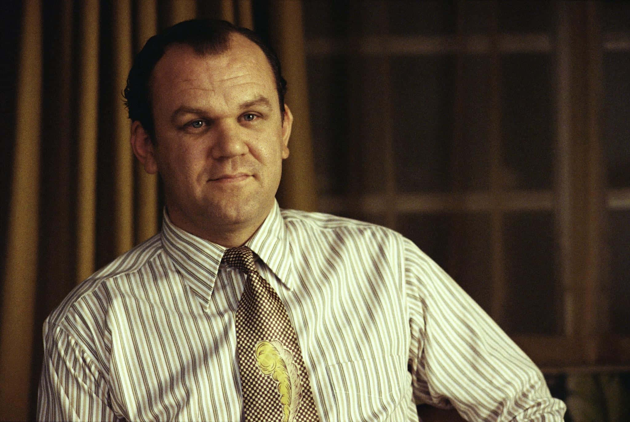 John C. Reilly 桌布 壁紙 — 一個穿著條紋襯衫和圖案領帶的男人，在一個光線昏暗、有木百葉窗的房間裡，帶著一絲微笑專注地凝視著。