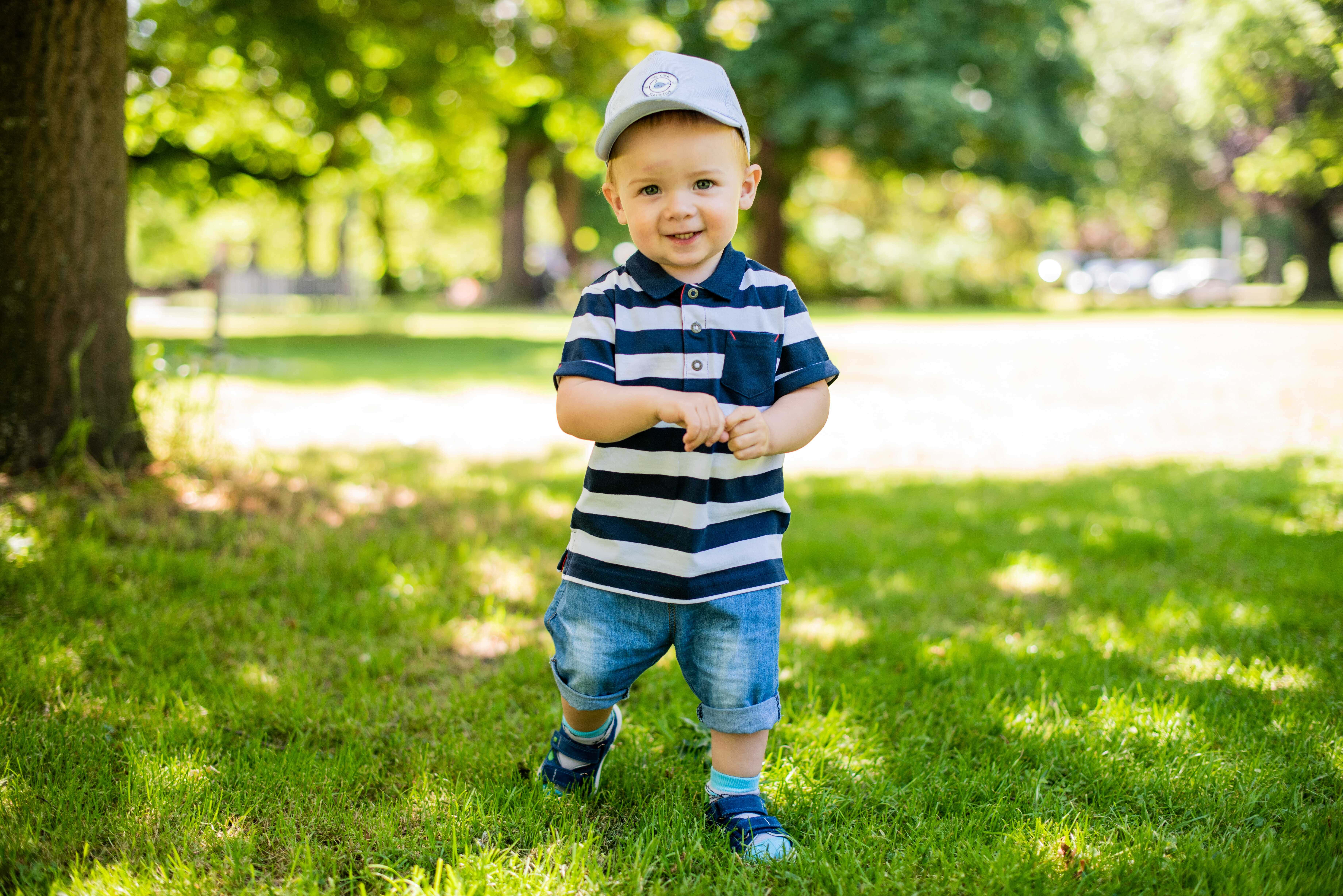 Baby Boy 桌布 壁紙 — 一個穿著條紋襯衫和帽子的快樂幼兒，在陽光明媚的草地上，站在枝葉茂密的樹下微笑。