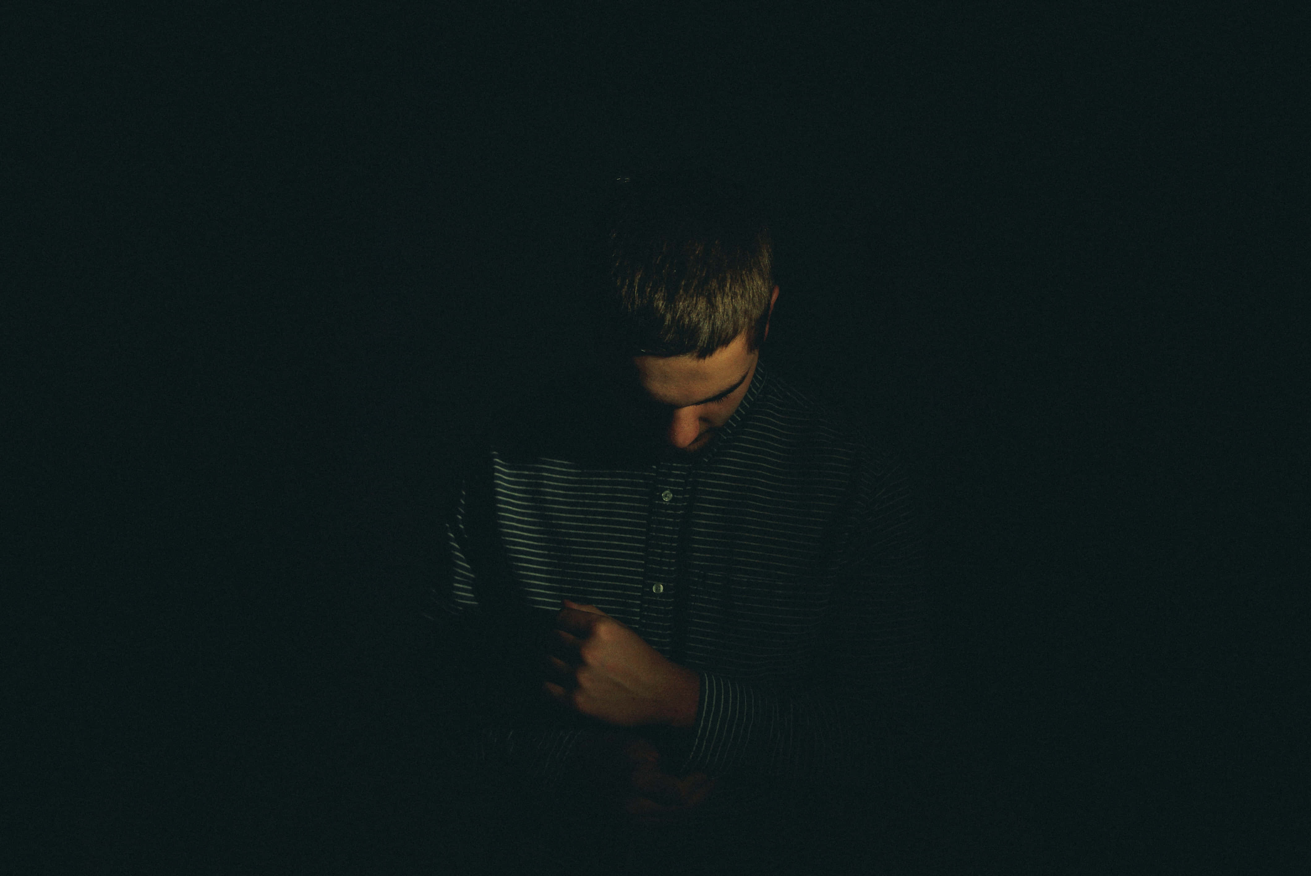Dark Emotional 桌布 壁紙 — 一個穿著條紋襯衫的人，在幾乎完全黑暗的環境中低著頭、雙手合十，被一道聚焦的光線照亮。