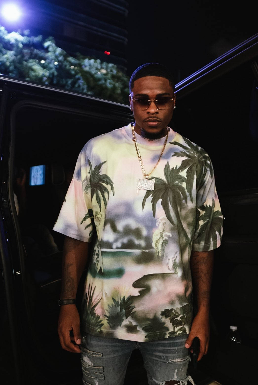Celebrity 桌布 壁紙 — 一個穿著棕櫚樹印花襯衫的男人在夜晚的城市燈光下自信地站在車旁。