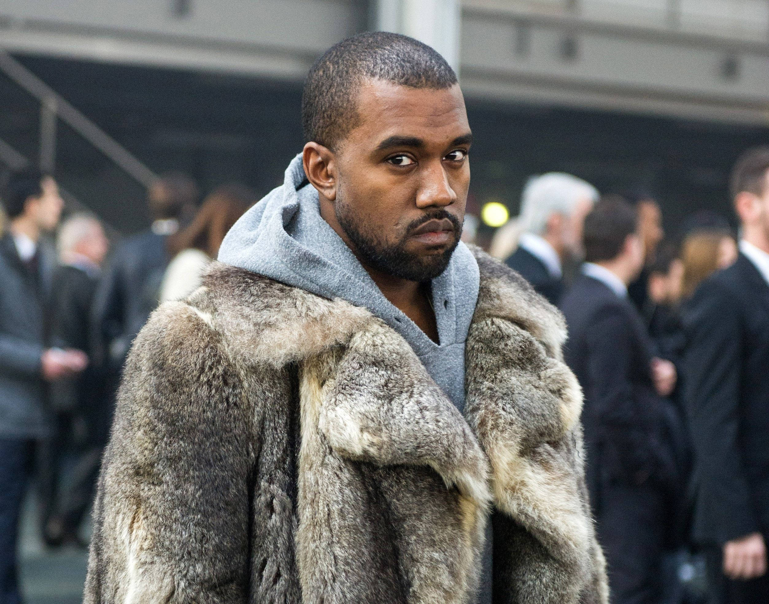 Kanye West Big Grey Fur 壁紙 — 一個穿著毛皮大衣和連帽衫的男人在模糊的人群中凝視著相機，身處城市環境。