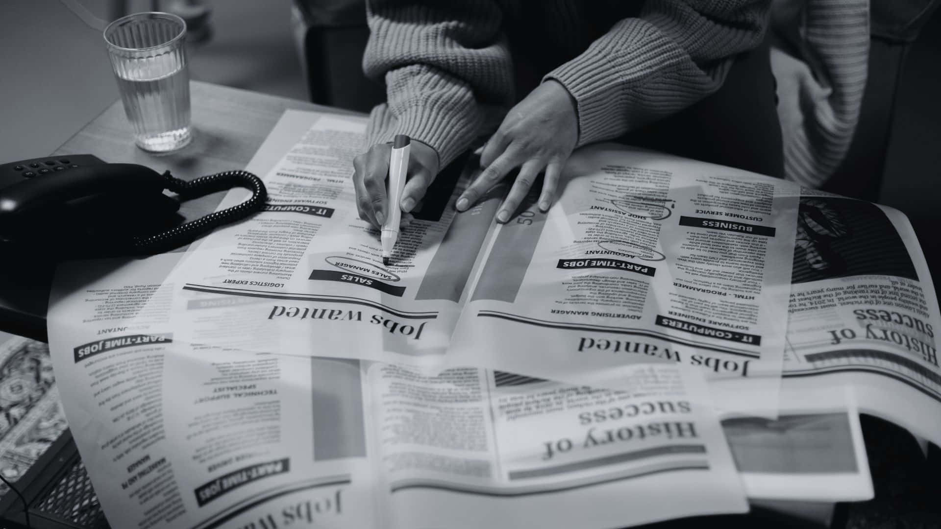 Black And White Newspaper 桌布 壁紙 — 一個穿著毛衣的人正在一份刊登招聘廣告的報紙上寫字，旁邊放著一杯水和一部電話。