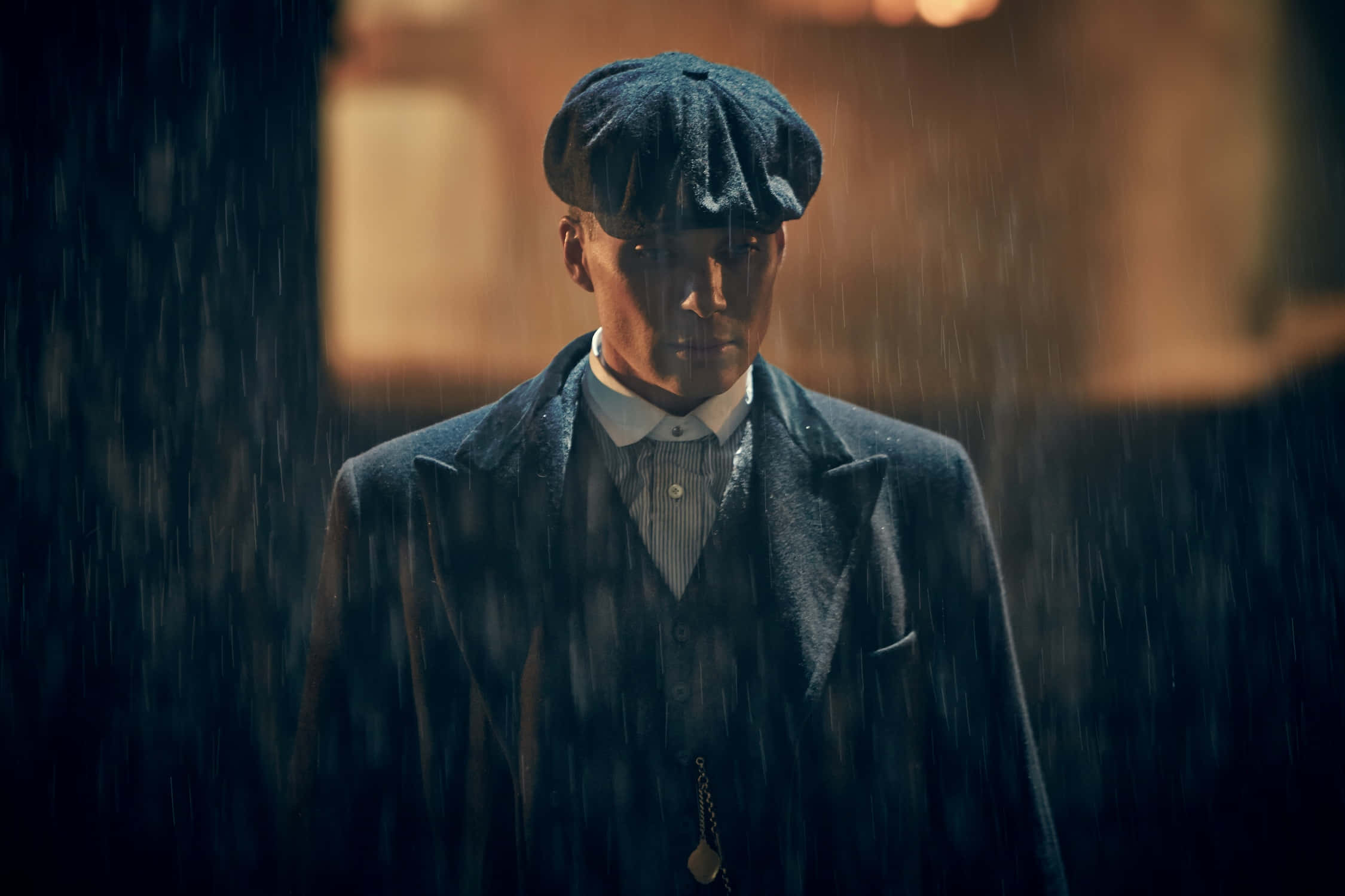 Peaky Blinders 桌布 壁紙 — 一個穿著深色外套和平頂帽的男人在溫暖的背光下莊嚴地站在大雨中。