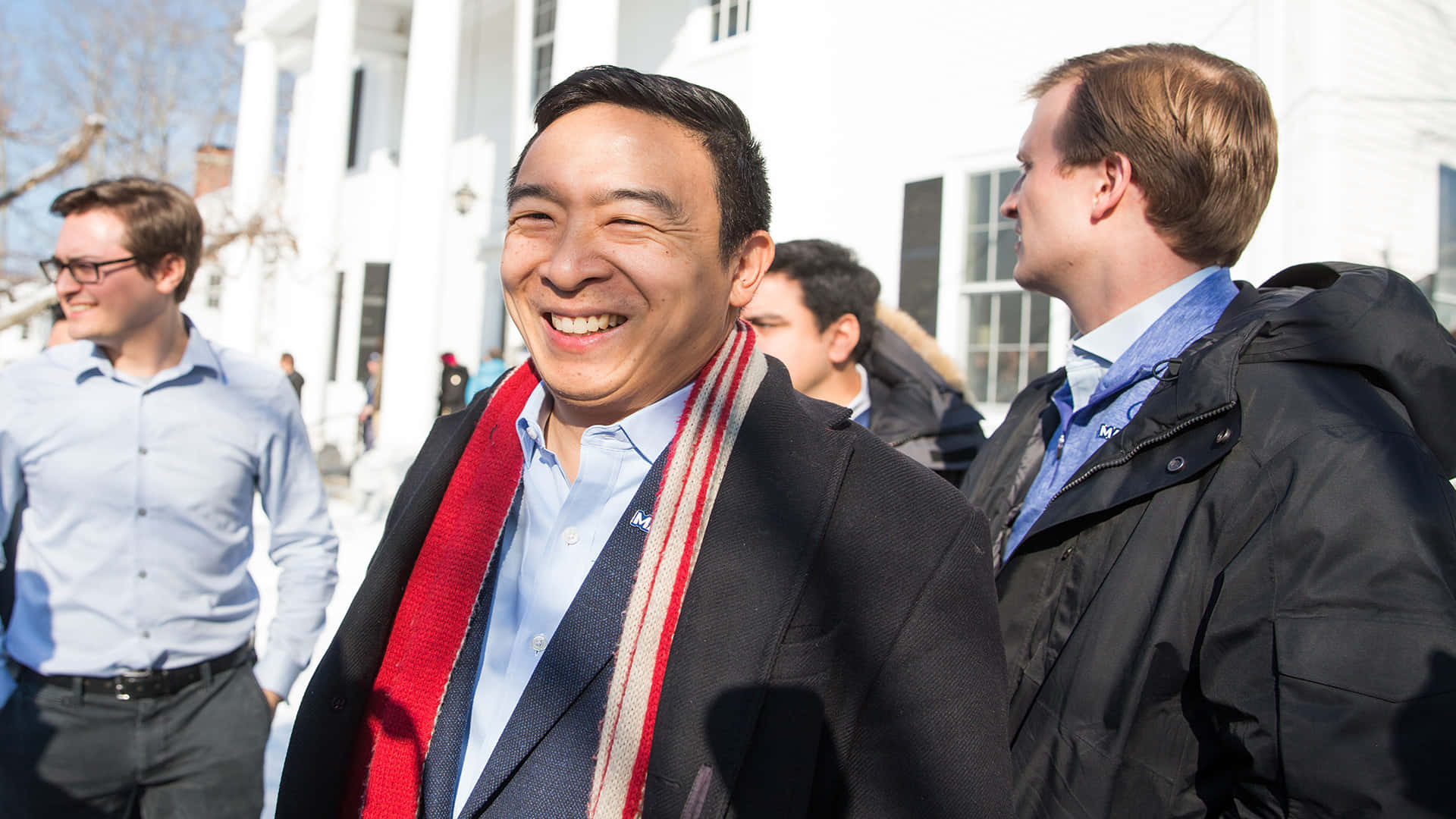Andrew Yang 桌布 壁紙 — 一個穿著深色外套和紅色圍巾的男人在晴朗的日子裡，與其他人在白色建築物附近開心地大笑。