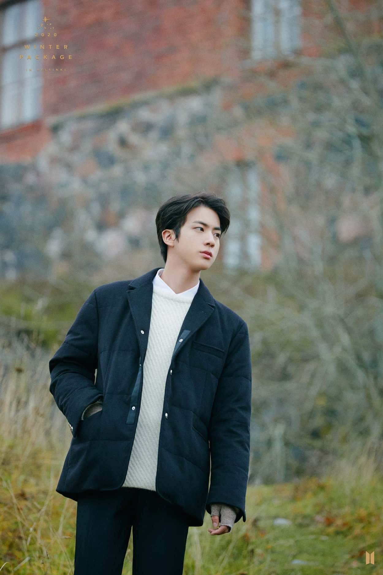 Jin 桌布 壁紙 — 一個穿著深色外套的年輕男子在柔和冬日陽光下，站在草地上，身後是磚牆，若有所思。