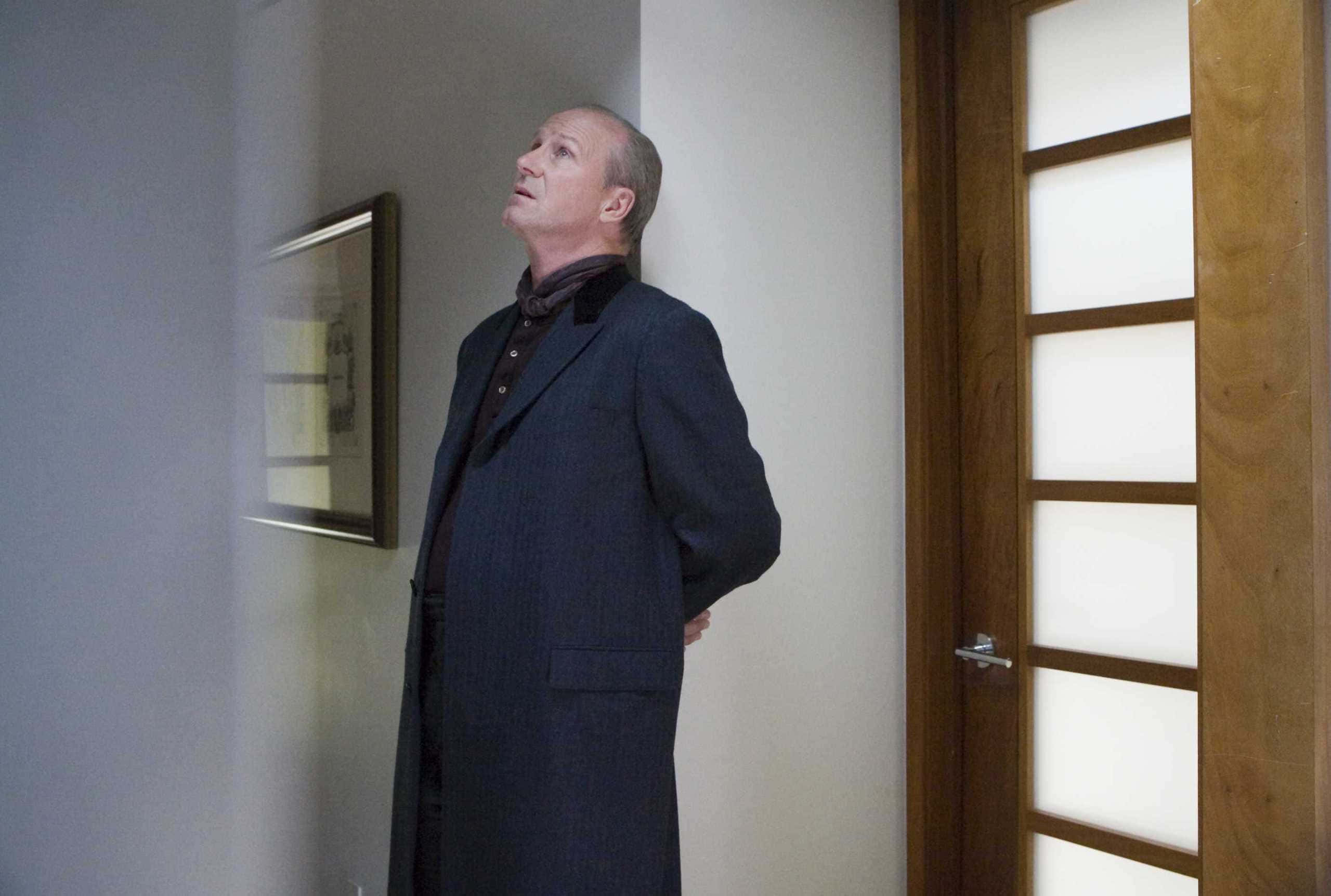 William Hurt 桌布 壁紙 — 一個穿著深色外套的男人站在走廊裡，抬頭仰望，表情若有所思，旁邊有一扇木門和一幅裱框藝術品。