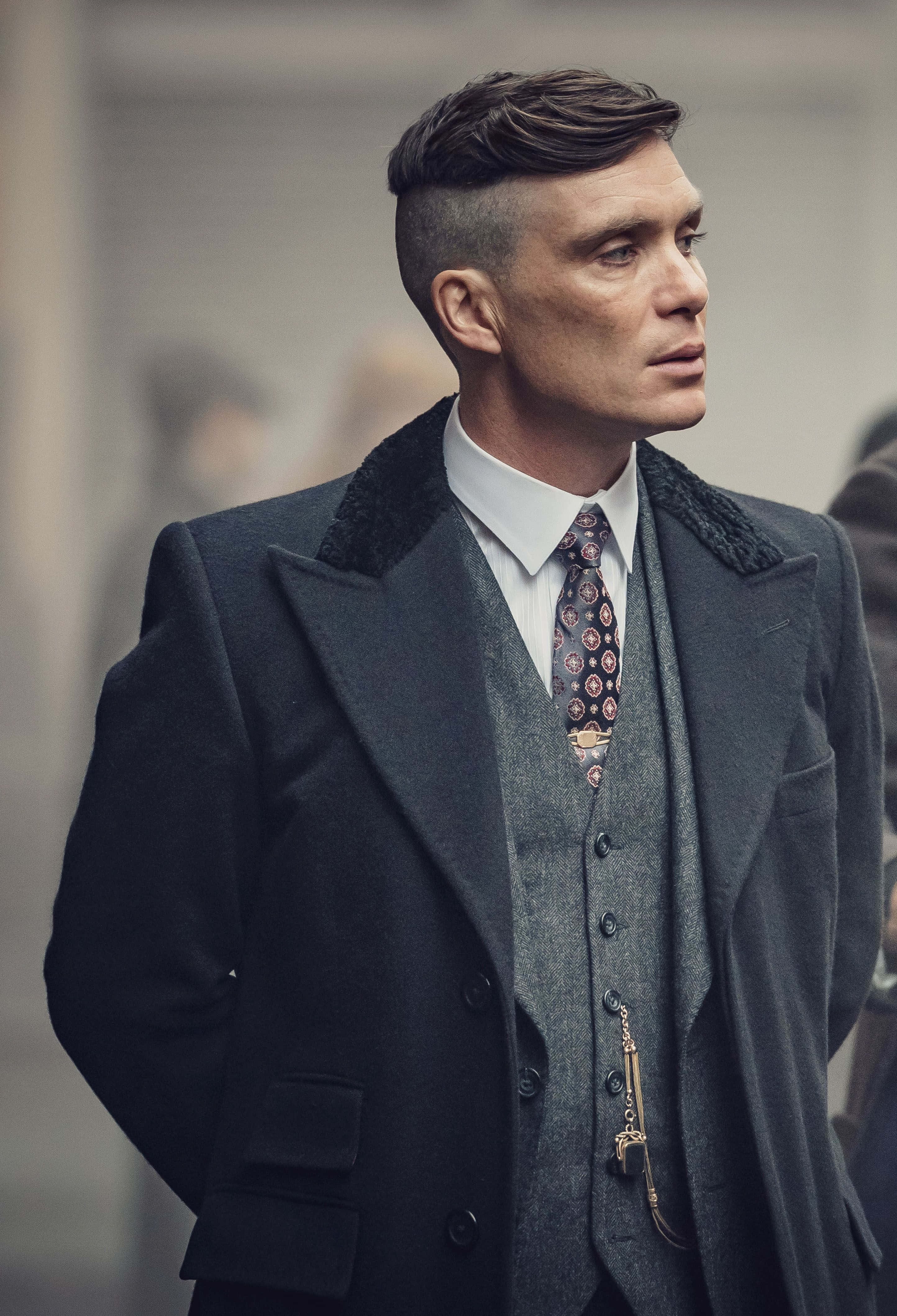 Peaky Blinders 桌布 壁紙 — 一個穿著深色羊毛外套和圖案領帶的男人，在模糊的城市背景下帶著嚴肅的表情站立。