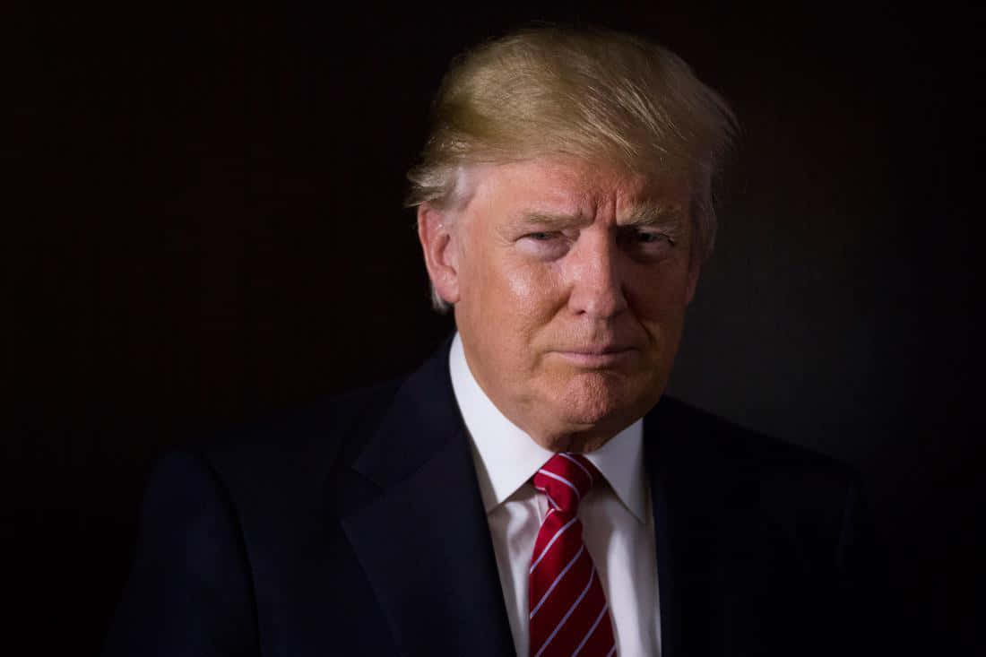 trump 桌布 壁紙 — 一個穿著深色西裝和紅色條紋領帶的男子在黑色背景前的嚴肅肖像。