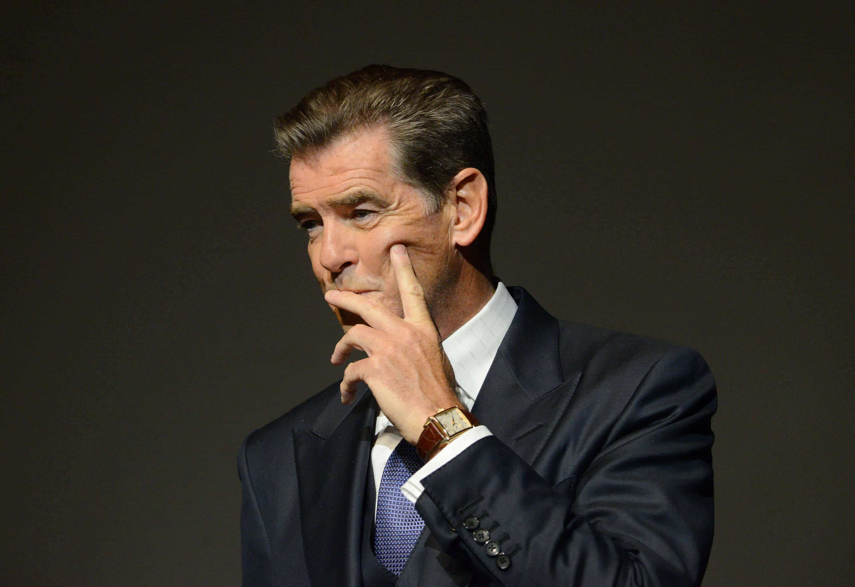 pierce brosnan 桌布 壁紙 — 一個穿著深色西裝和領帶的男人，在純色深色背景上用手托著下巴。