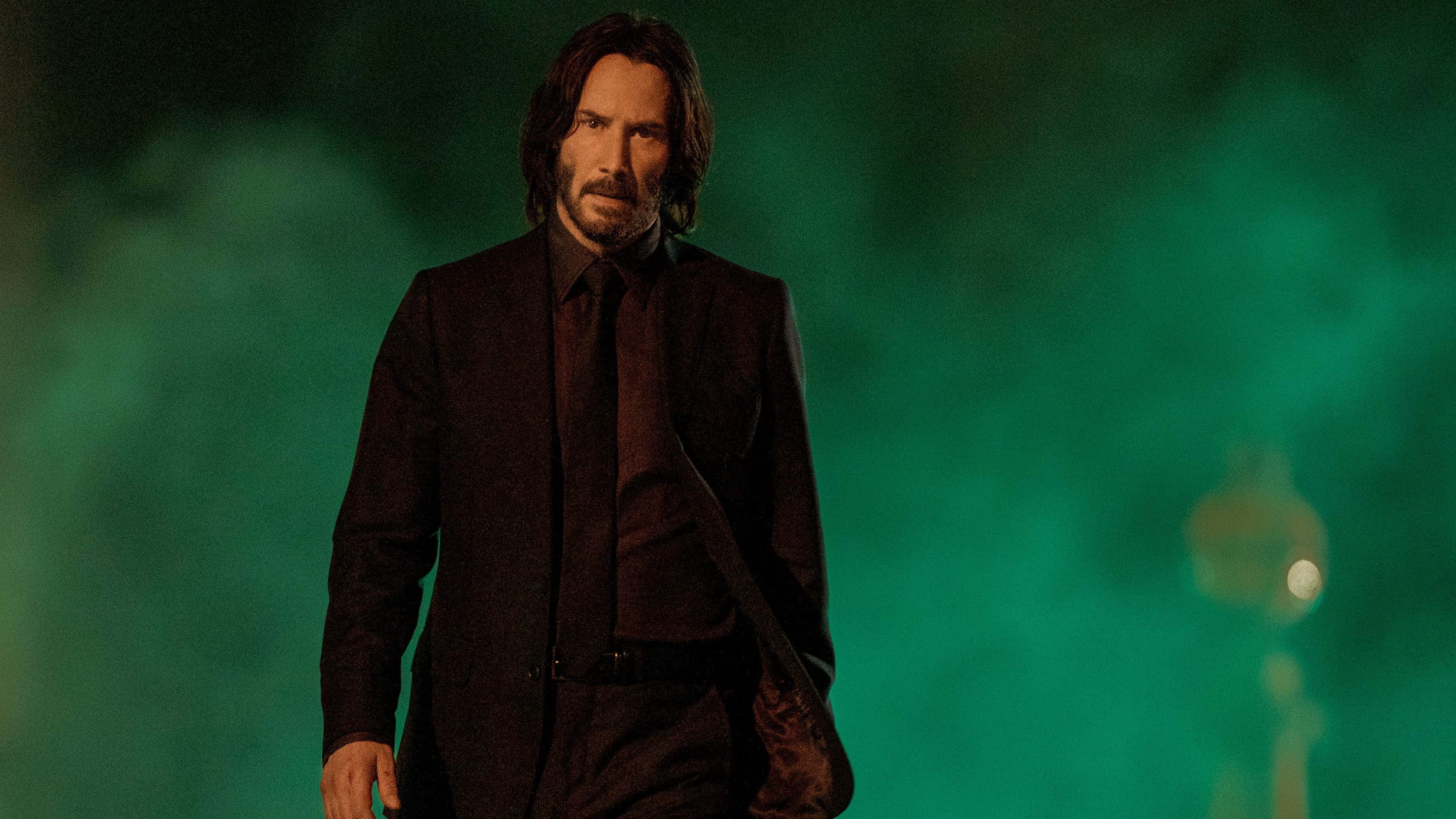 Keanu Reeves 桌布 壁紙 — 一個穿著深色西裝的男人，在朦朧的綠色燈光背景和模糊的路燈下莊嚴地站立。