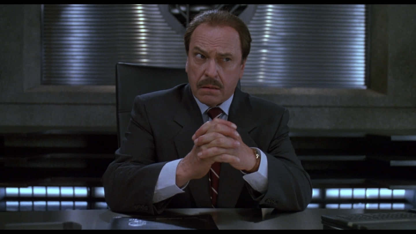 Rip Torn 桌布 壁紙 — 一個穿著深色西裝的男人坐在辦公桌前，雙手緊握，在燈光昏暗的未來辦公室裡顯得憂心忡忡。