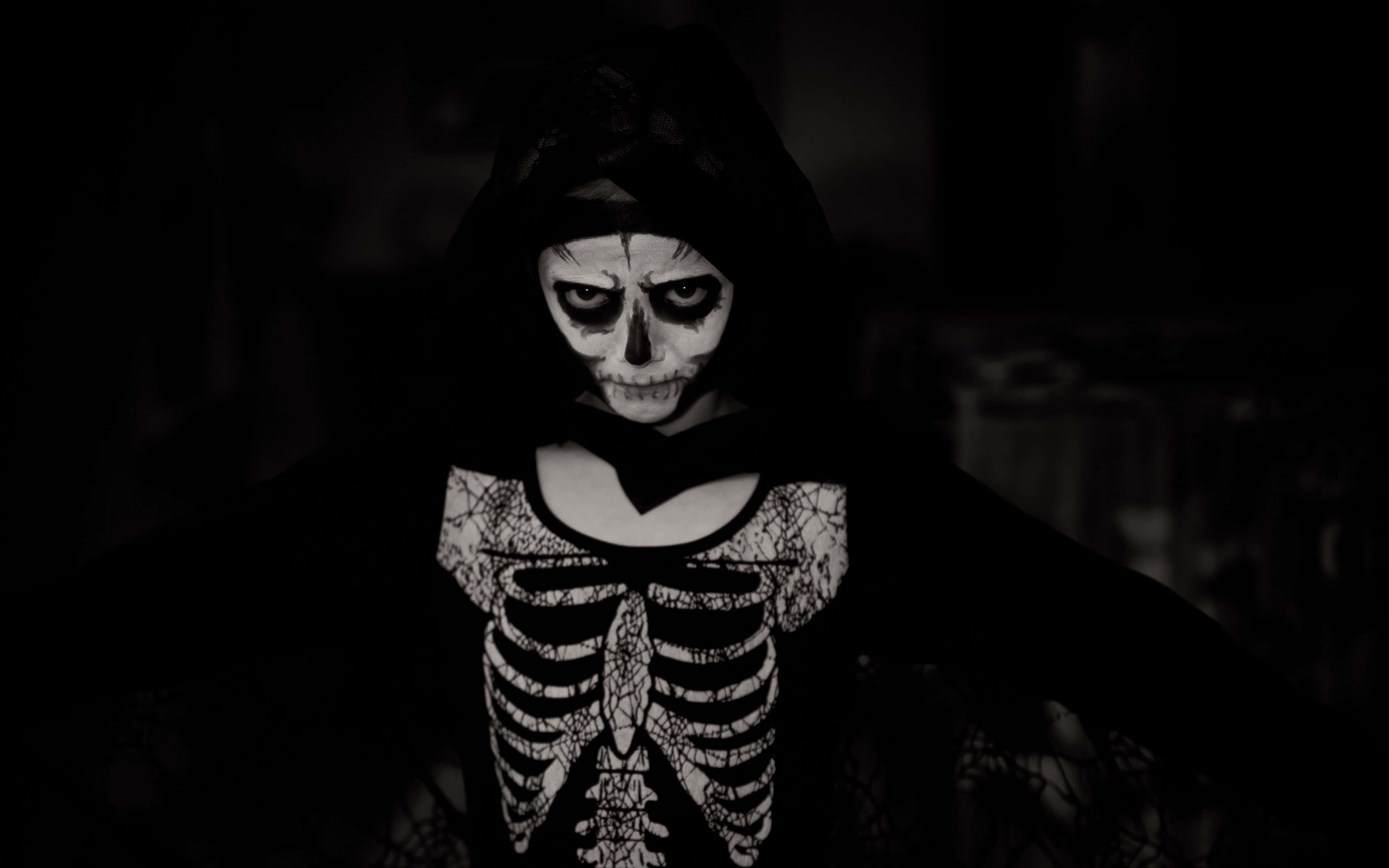 Halloween Skeleton 桌布 壁紙 — 一個穿著深色連帽服裝、化著骷髏妝、穿著骷髏印花襯衫的人站在陰暗的朦朧中。