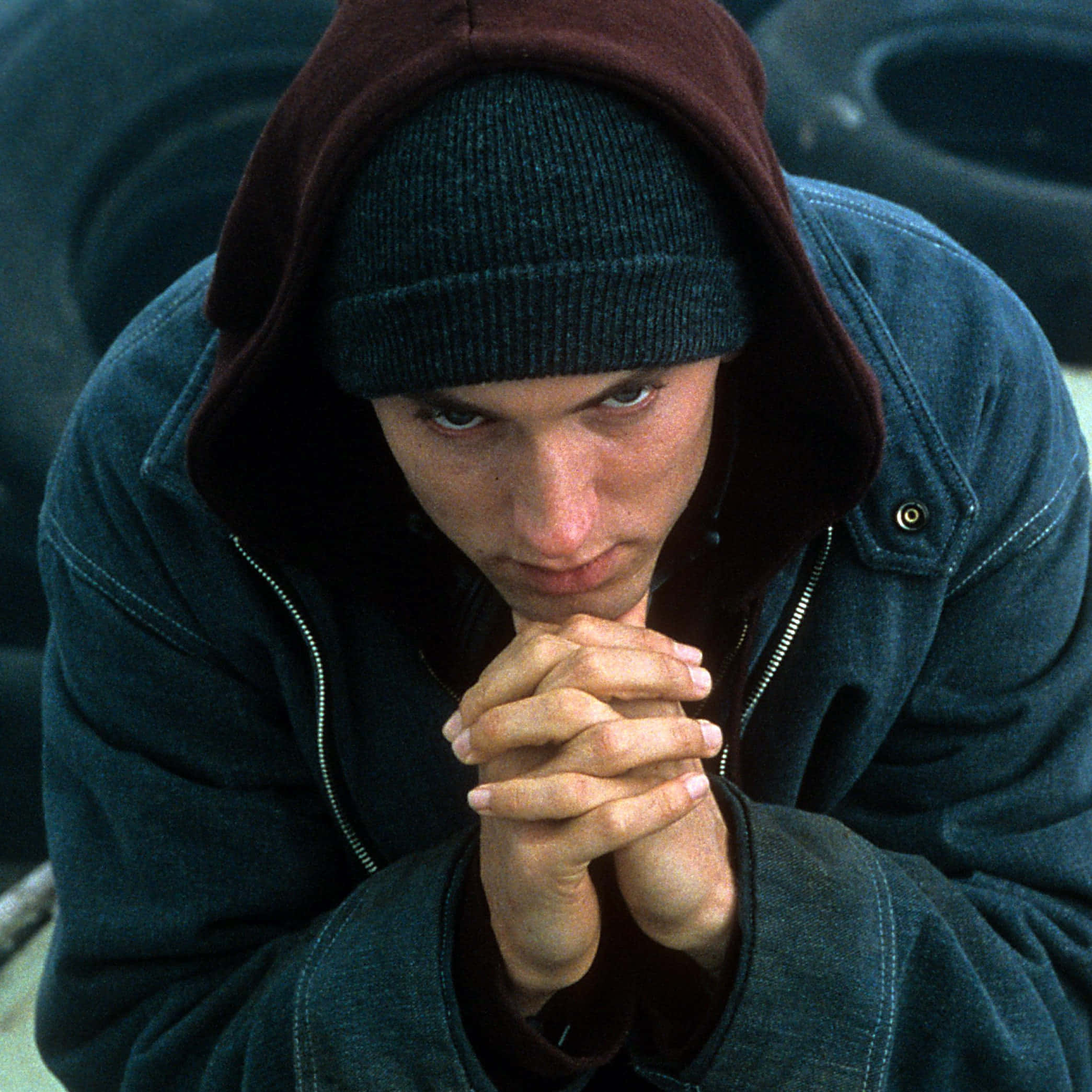 Eminem 桌布 壁紙 — 一個穿著深色連帽衫和冷帽的年輕男子，雙手緊握，在堆疊輪胎的背景前凝視著。