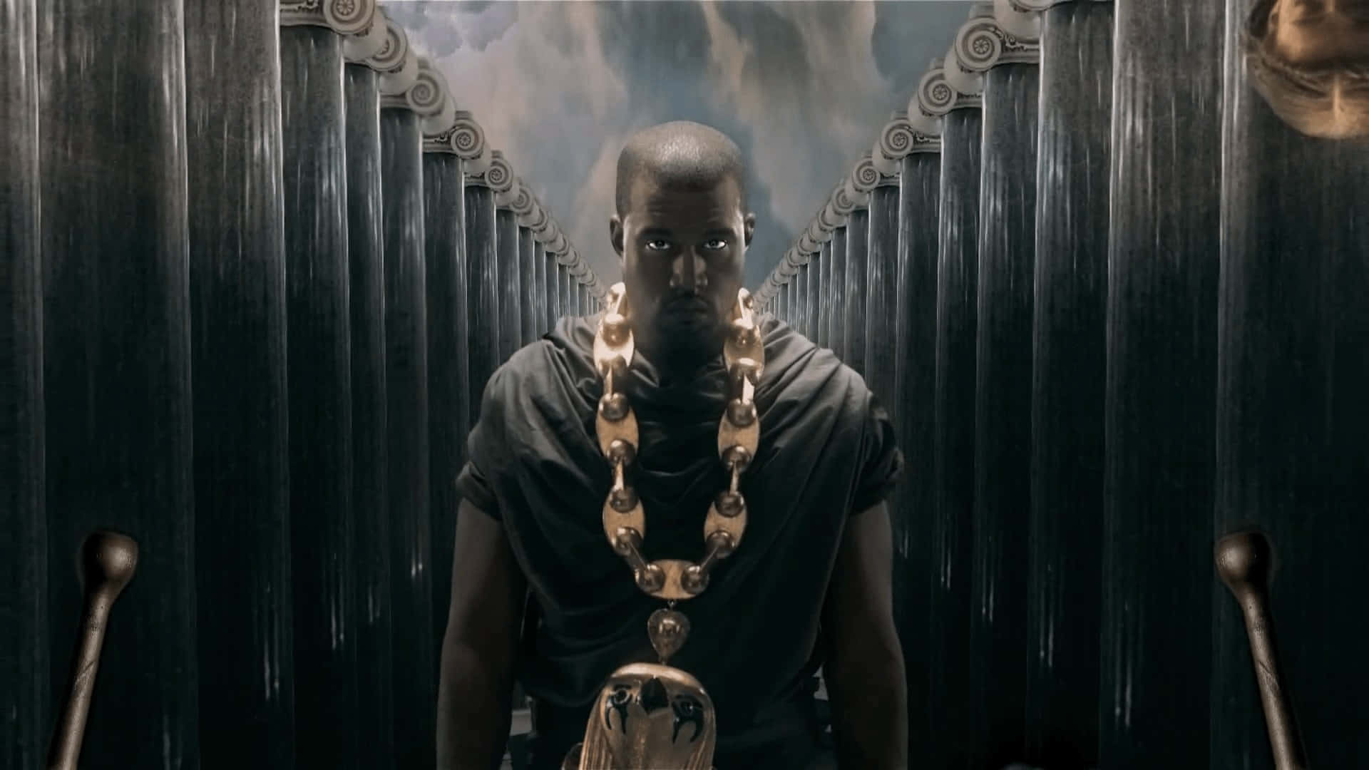 Kanye 桌布 壁紙 — 一個穿著深色長袍、戴著大金鏈的男人，在暴風雨的天空下，莊嚴地站在高聳華麗的柱子之間。