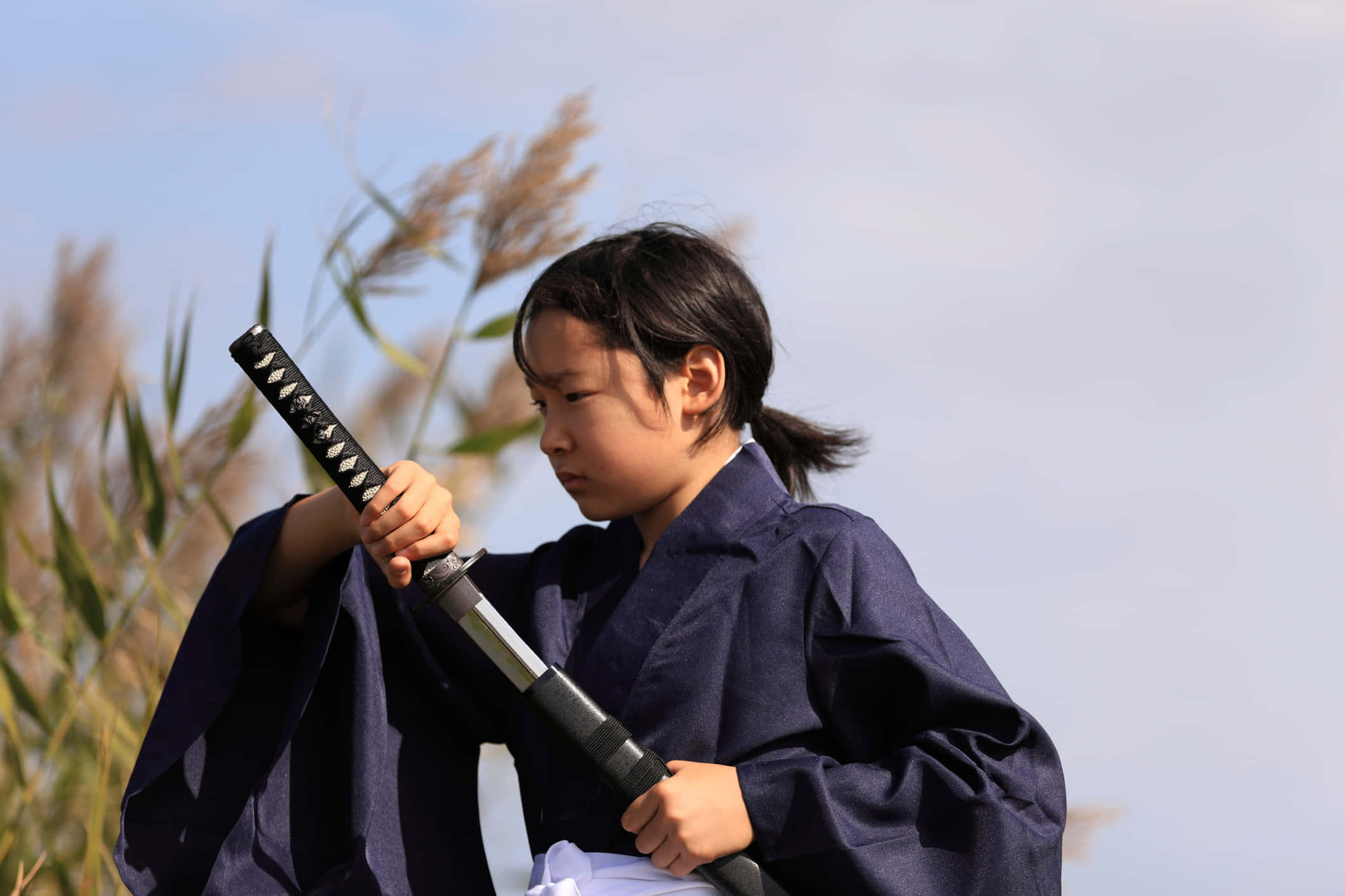 Samurai Sword 桌布 壁紙 — 一個穿著深藍色和服的少女，在高高的蘆葦和柔和天空的背景下，專注地握著一把刀。
