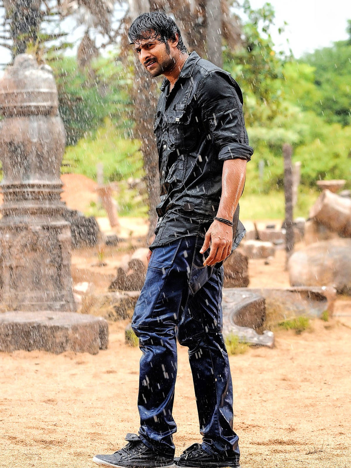 Prabhas Mirchi 桌布 壁紙 — 一個穿著濕透黑色襯衫和牛仔褲的男人站在雨中，周圍是古老的石頭廢墟，被綠意環繞。