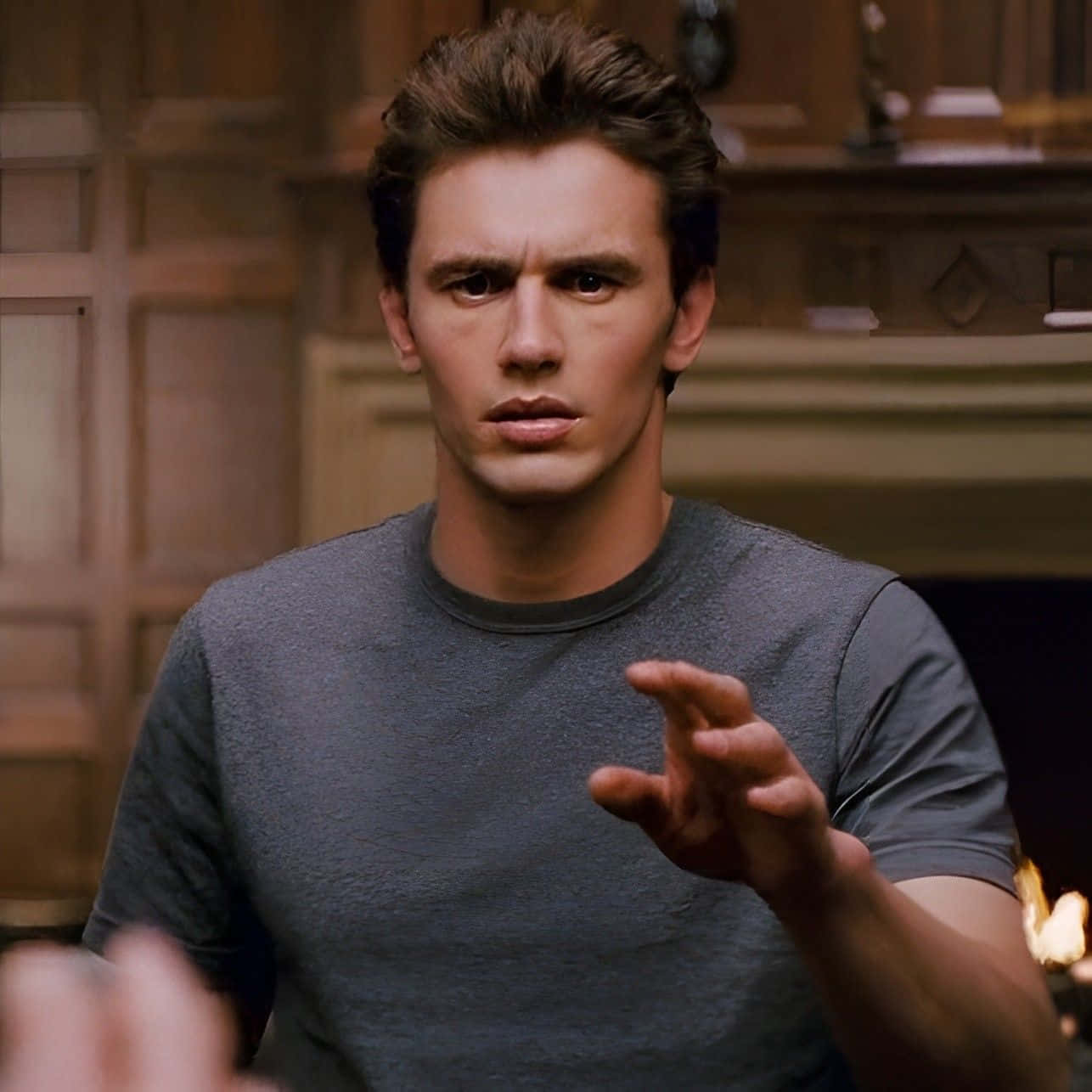 Harry Osborn 桌布 壁紙 — 一個穿著灰色T恤的男人在一個溫暖燈光的木鑲板房間裡，壁爐旁急切地做手勢。