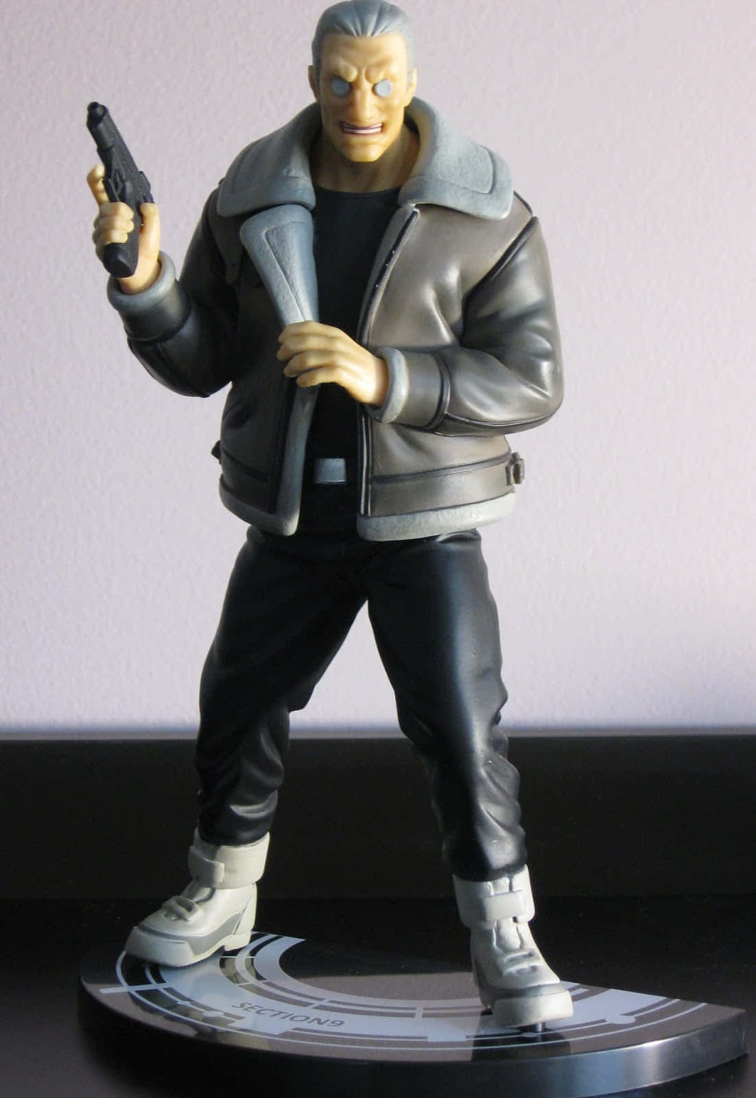 Ghost In The Shell Batou 桌布 壁紙 — 一個穿著灰色外套、手持手槍的男性動漫人物模型，細節精緻，站立在圓形底座上，背景是素牆。