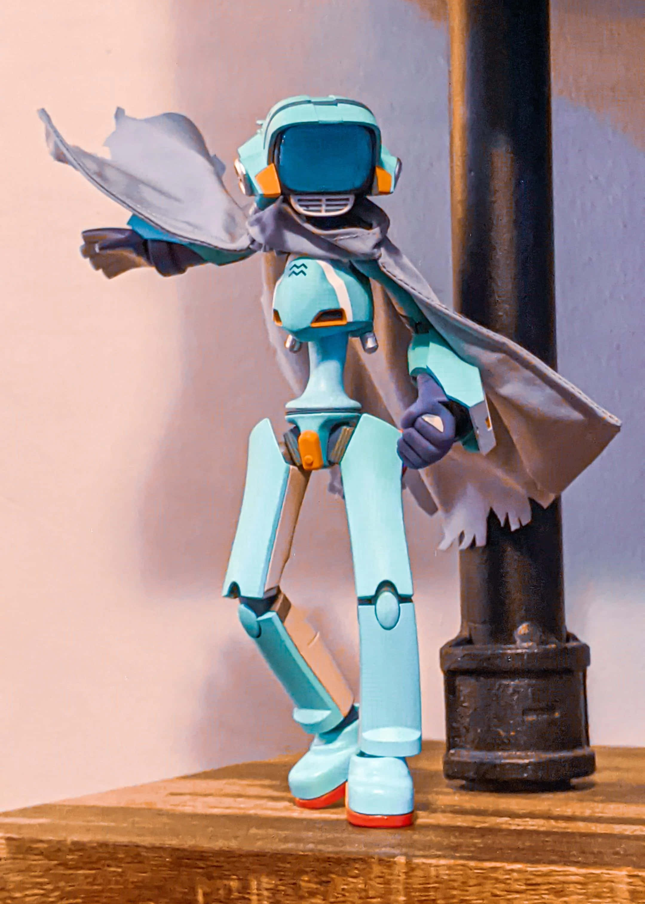 Flcl Canti 桌布 壁紙 — 一個穿著灰色斗篷的藍綠色機械人公仔，英雄式地站在木質表面上，旁邊有一根黑色柱子。