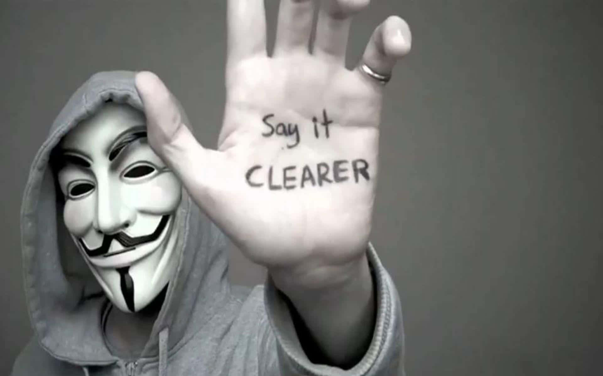 Anonymous 桌布 壁紙 — 一個穿著灰色連帽衫、戴著蓋伊·福克斯面具的人舉起一隻手，手掌上寫著「Say it CLEARER」。