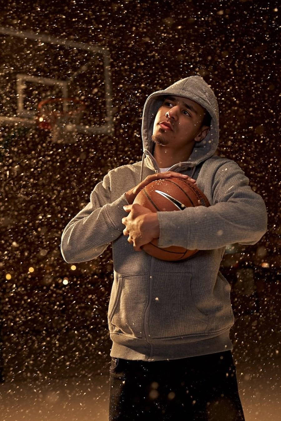 J Cole《The Warm Up》 壁紙 — 一個穿著灰色連帽衫的年輕人在飄雪中拿著籃球，背景有一個模糊的籃球框。