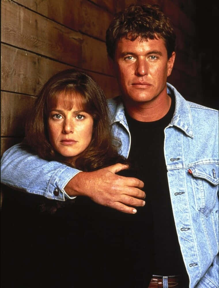 Tom Berenger 桌布 壁紙 — 一個穿著牛仔外套的男人在木牆前擁抱一個女人，形成一幅戲劇性而親密的肖像。