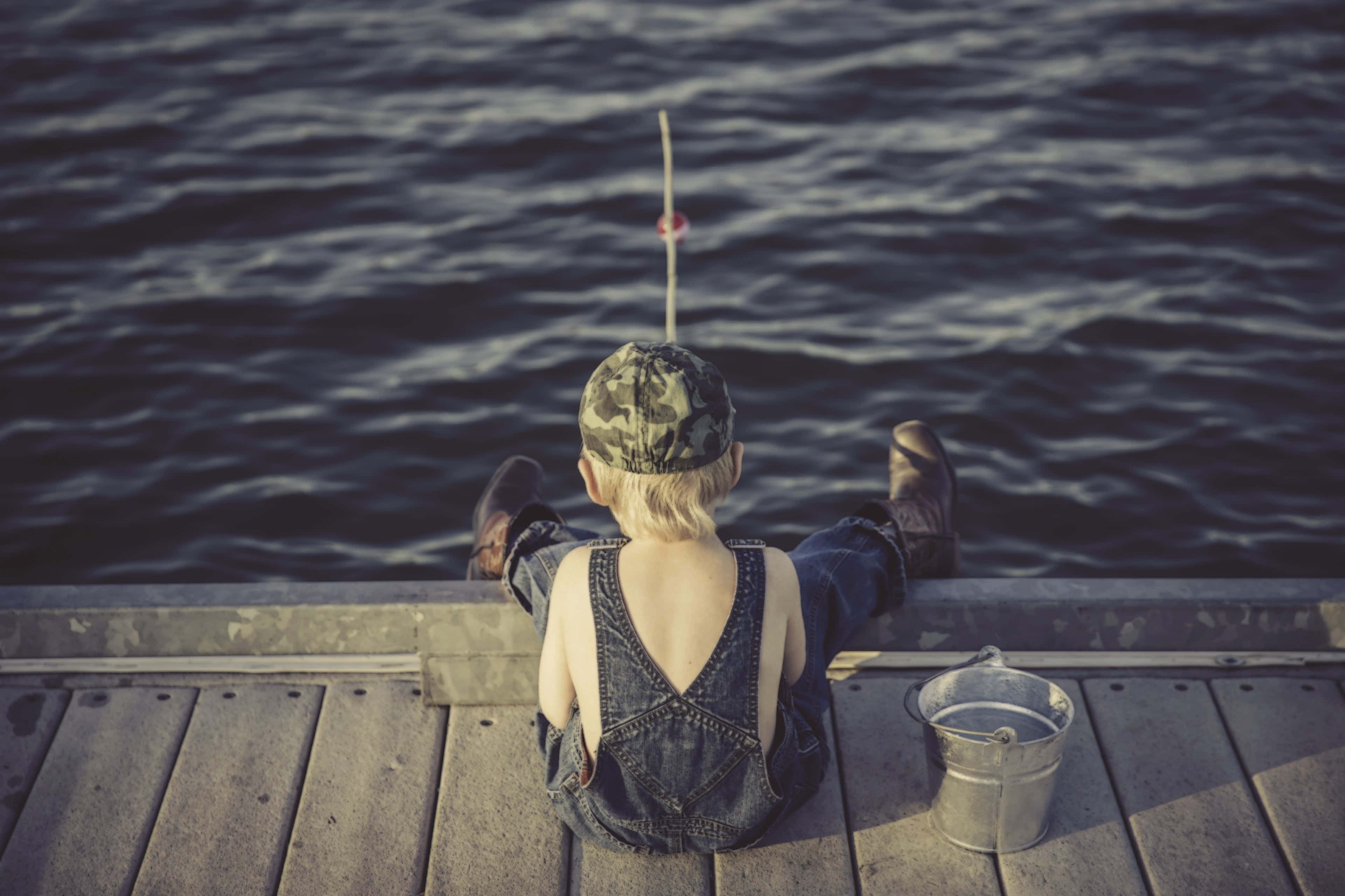 Fishing Rod 桌布 壁紙 — 一個穿著牛仔工裝褲和迷彩帽的小孩坐在木碼頭上釣魚，浮標在平靜的水面上晃動。