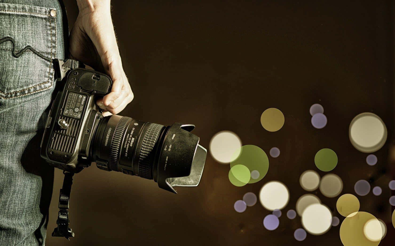 Dslr Camera 桌布 壁紙 — 一個穿著牛仔褲的人手持一部DSLR相機，在黑暗背景下發出色彩繽紛的散景光。