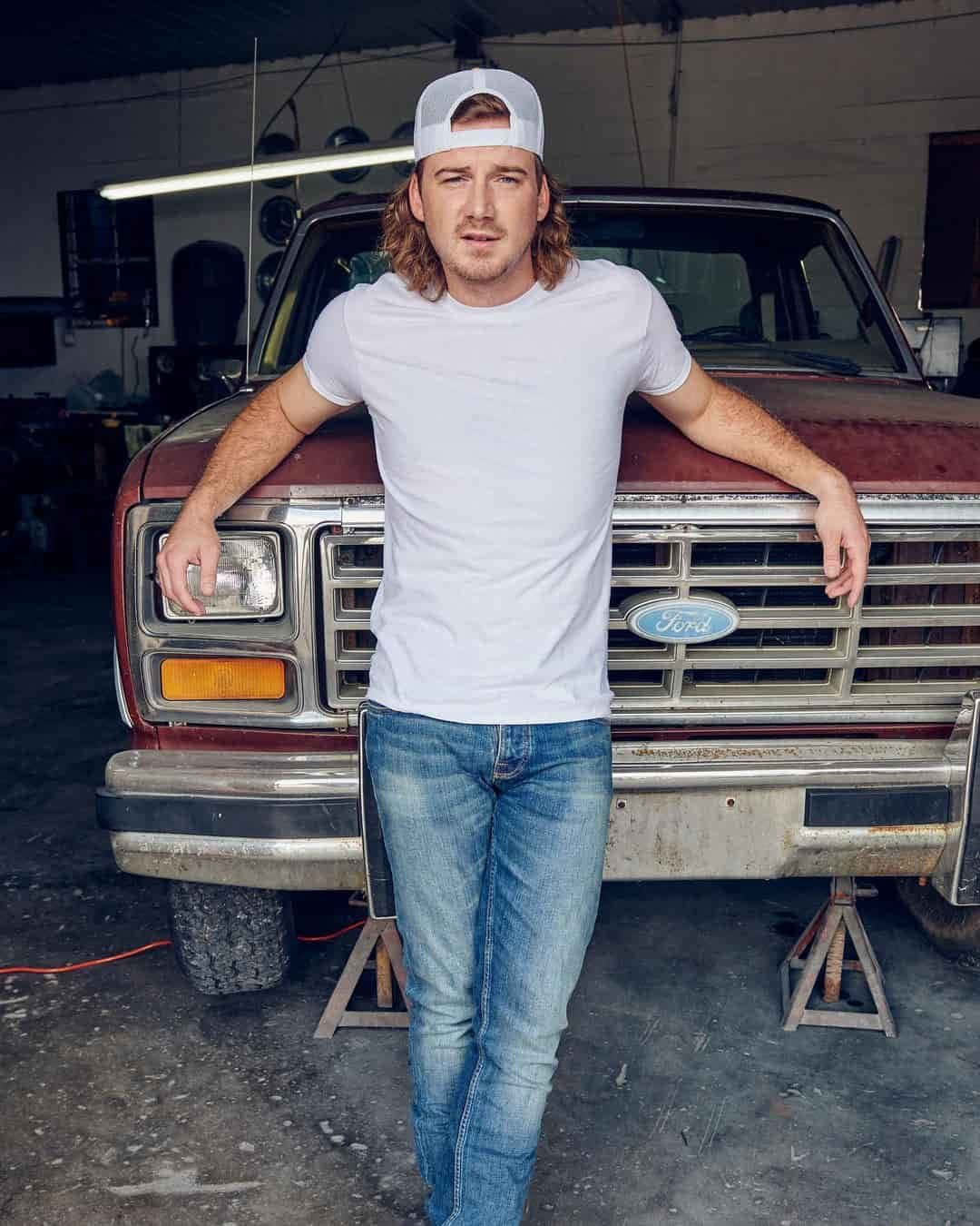 Morgan Wallen 桌布 壁紙 — 一個穿著白色T恤和牛仔褲的男人，在燈光昏暗的車庫裡，隨意地靠在一輛生鏽的紅色福特卡車前。