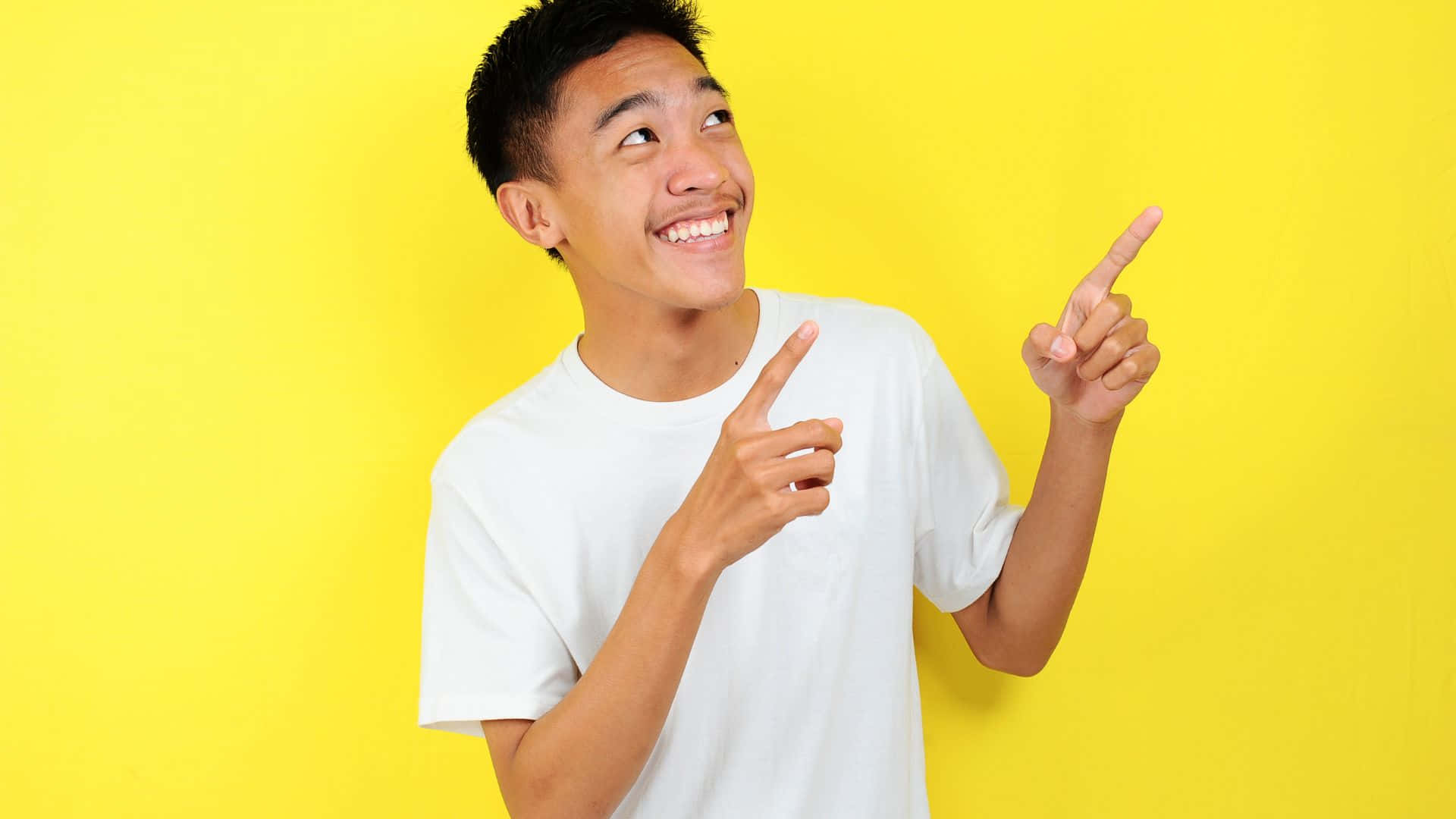 Boy 桌布 壁紙 — 一個穿著白色T恤的開朗年輕男子在明亮的黃色背景前微笑並向上指。