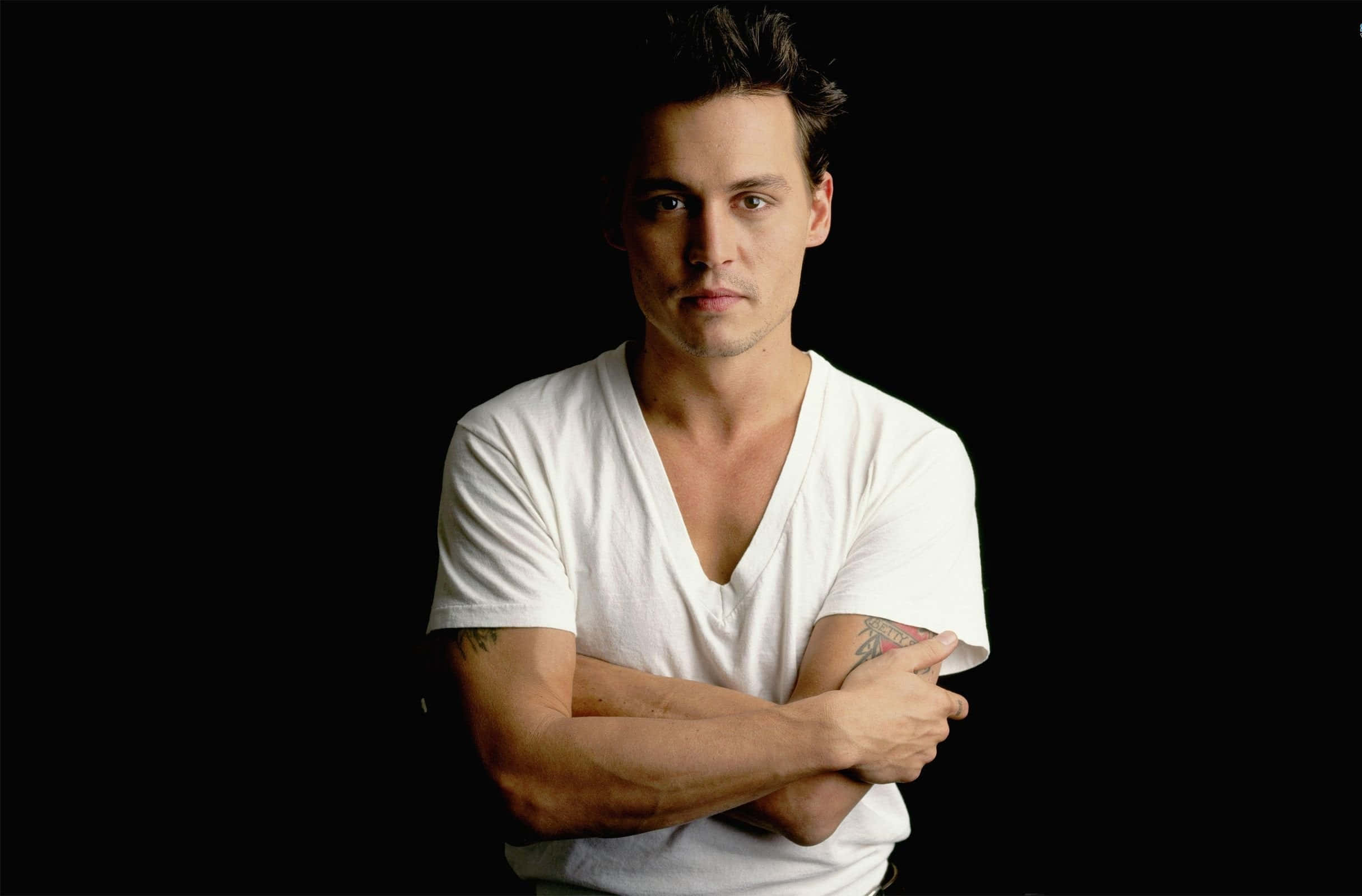 Johnny Depp 桌布 壁紙 — 一個穿著白色V領T恤的男人，雙臂交叉，站在純黑色背景前，眼神直接而強烈。