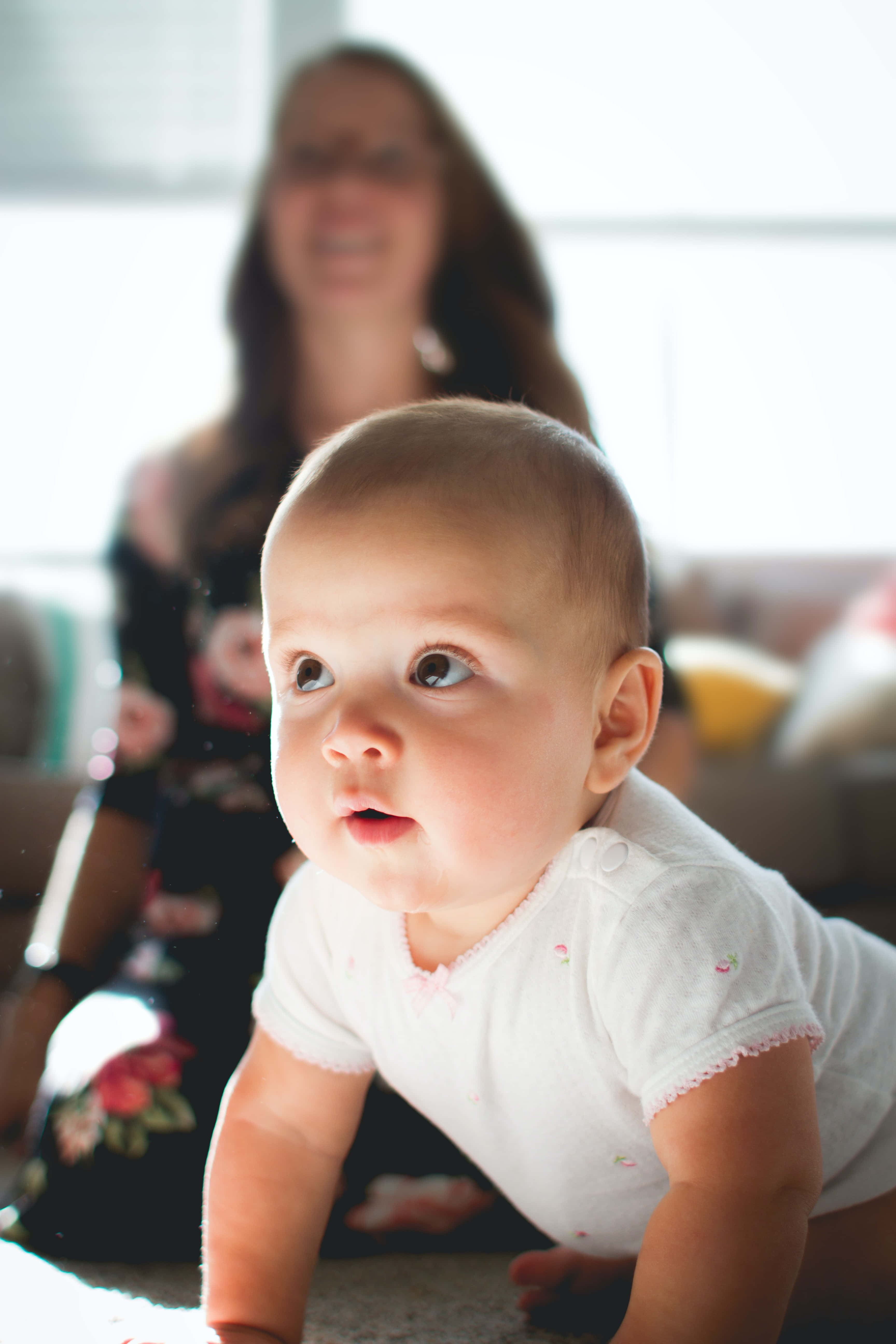 Baby Boy 桌布 壁紙 — 一個穿著白色連身衣的嬰兒在地毯上爬行，背景中一位微笑的女士模糊地坐著。