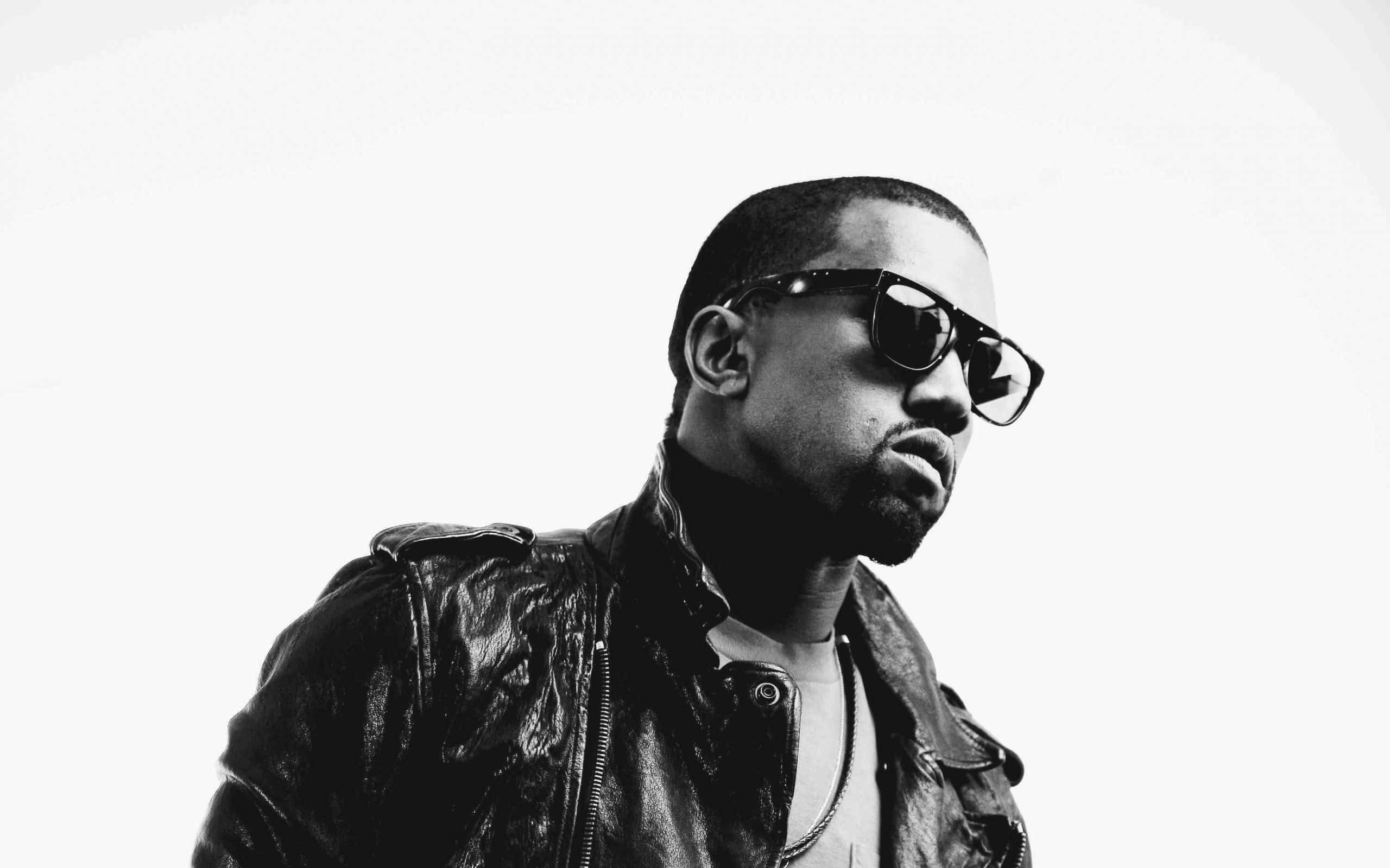 Kanye 桌布 壁紙 — 一個穿著皮褸和太陽眼鏡的男人，在純白背景前以高對比黑白風格凝視。