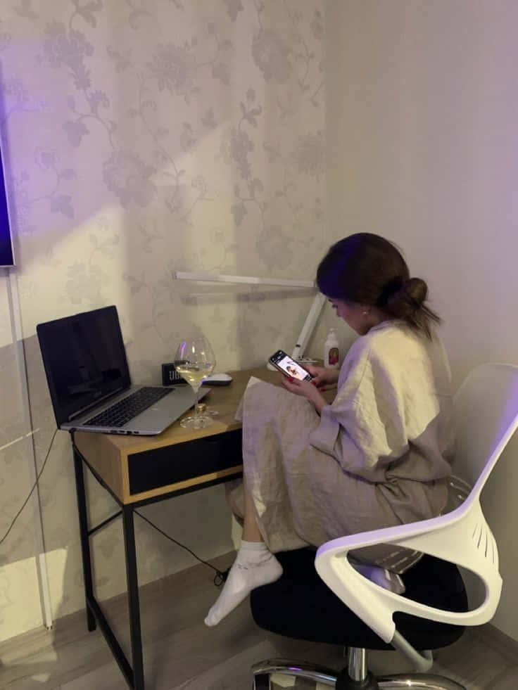 Studying 桌布 壁紙 — 一個穿著睡袍的人坐在一張書桌前，房間燈光柔和，牆紙是花卉圖案，旁邊有手提電腦和酒杯，正在使用電話。