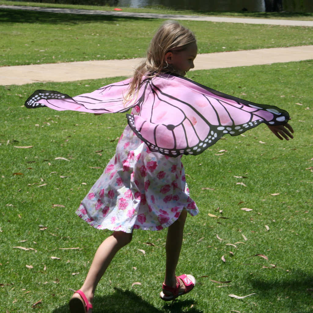 Butterfly Wing Dress 桌布 壁紙 — 一個穿著碎花裙的小女孩，戴著粉紅色帝王蝶翅膀，在陽光明媚的草地上奔跑。