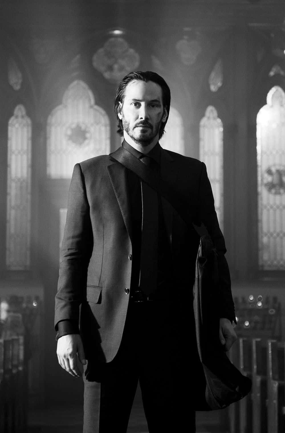 Keanu Reeves 桌布 壁紙 — 一個穿著筆挺西裝的男人莊嚴地站在光線昏暗的大教堂裡，彩色玻璃窗在他身後投射出柔和的光芒。