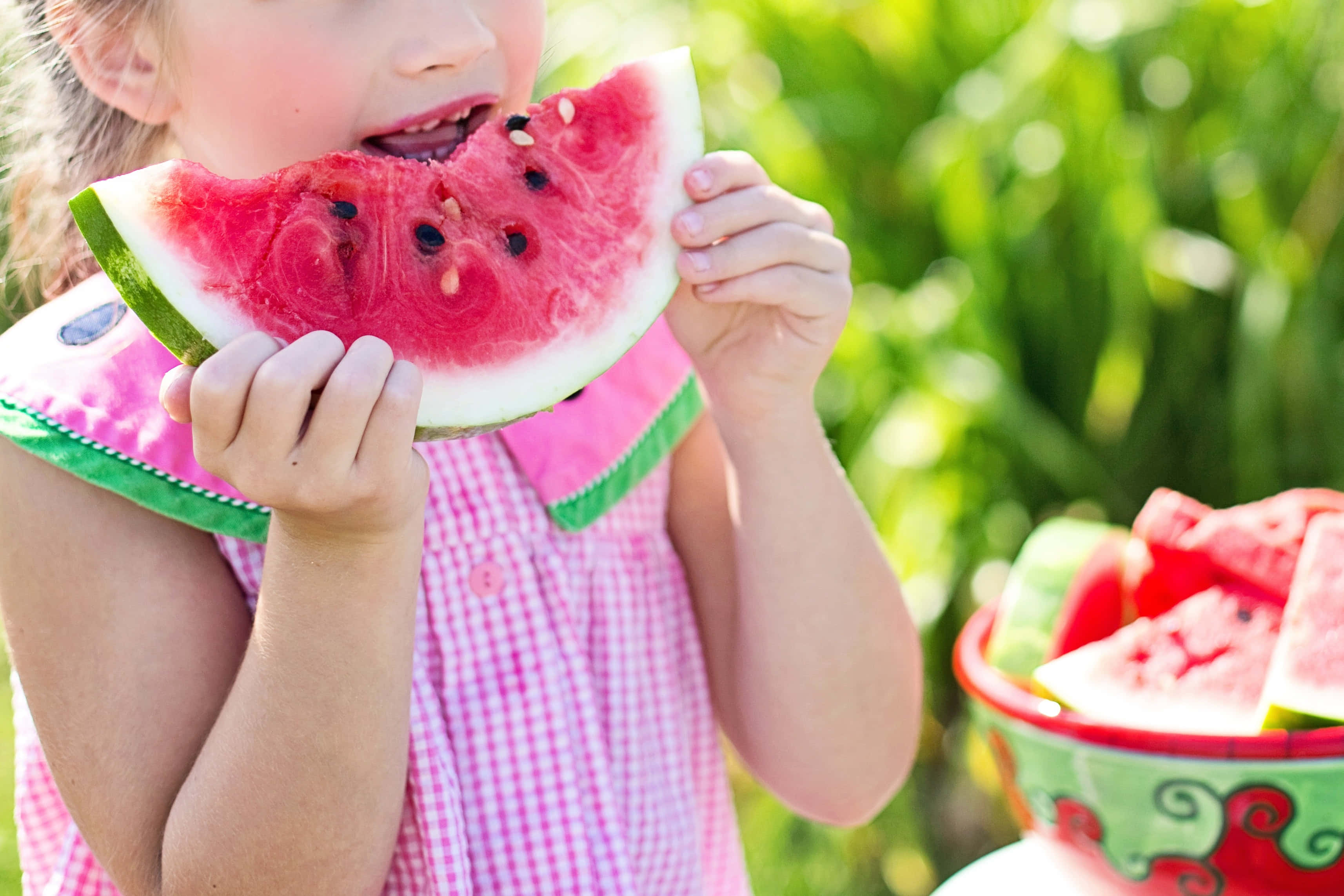 Summer Fruits 桌布 壁紙 — 一個穿著粉紅色格仔裙的小女孩，喺戶外開心咁咬住一塊多汁嘅西瓜。