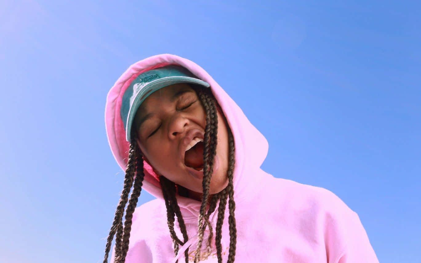 Lil Uzi Vert 桌布 壁紙 — 一個穿著粉紅色連帽衫和青綠色帽子的年輕人，在清澈的藍天下打著大哈欠。